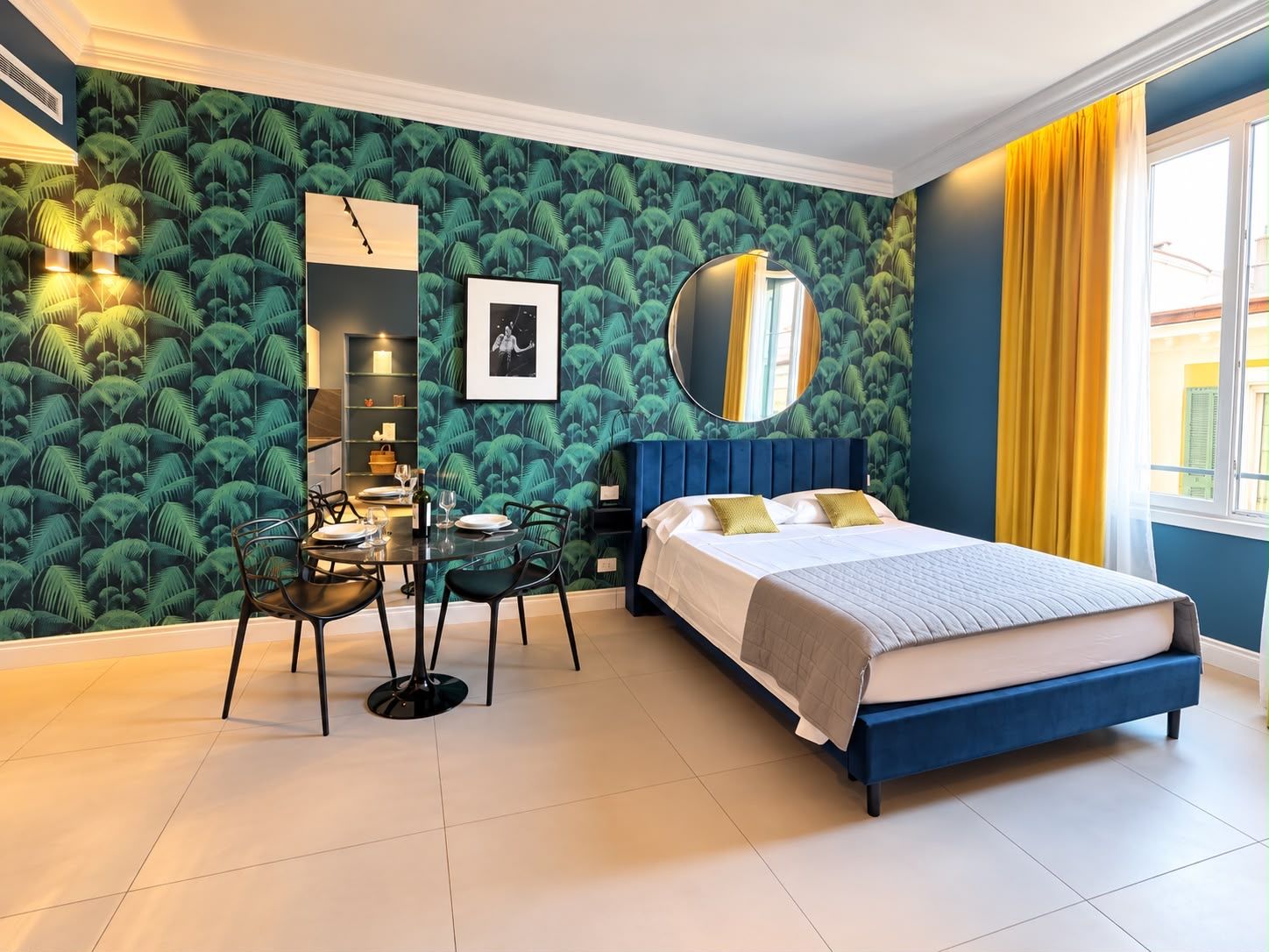 A bedroom with a bed, a dining table with chairs, a mirror, and a wall with a leafy pattern.