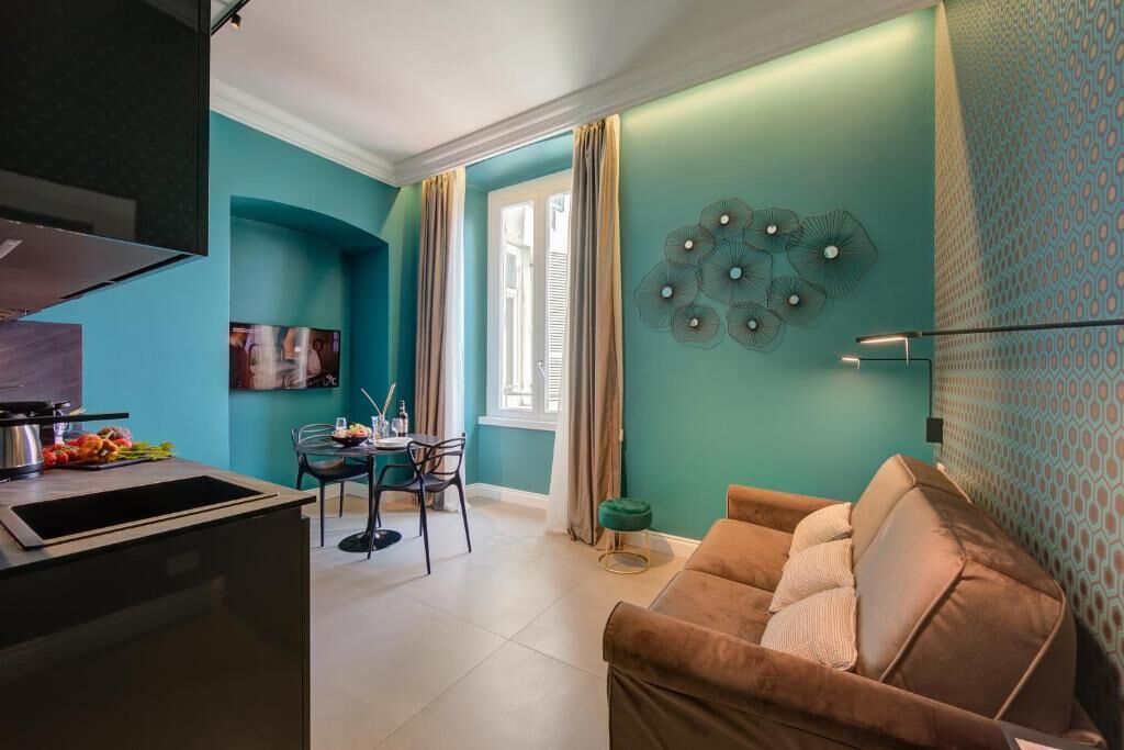 A modern living room with teal walls, a brown sofa, a dining area with a table and chairs, and a kitchen with a sink and stove.