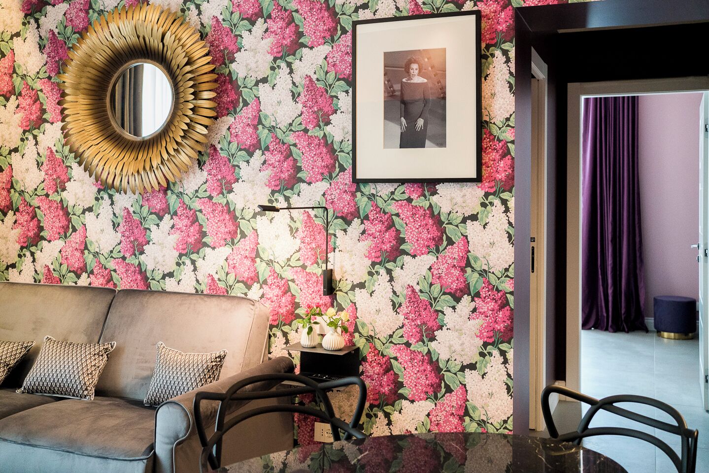Un salon avec un papier peint à motifs floraux, un miroir rond, un tableau encadré, un canapé avec des coussins à motifs et une table d’appoint avec des vases.