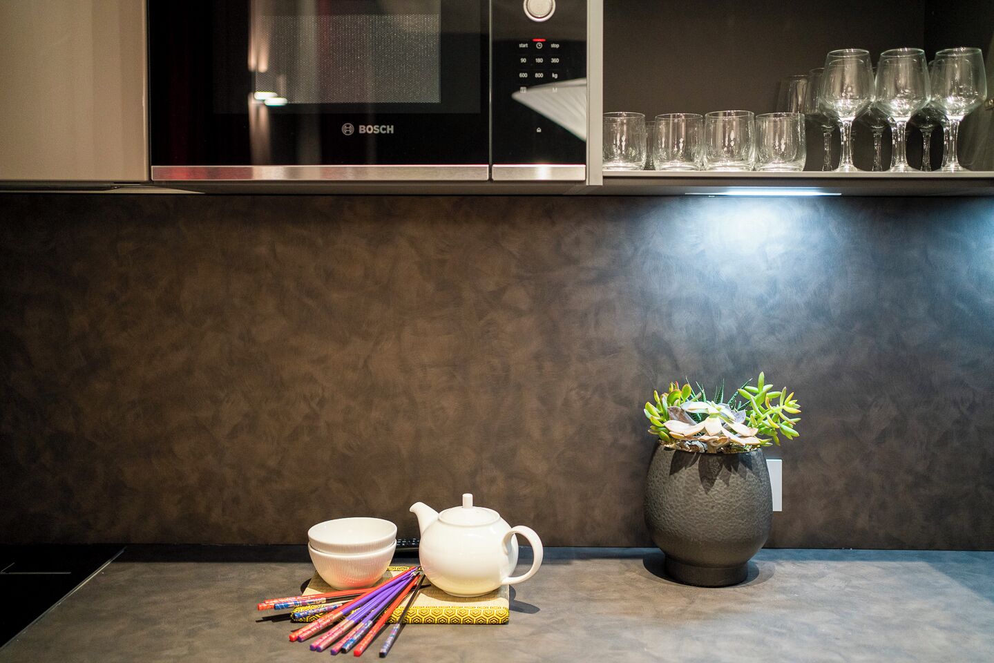 A kitchen with a Bosch microwave, a potted plant, a white teapot, a white bowl, and colorful chopsticks on a countertop.