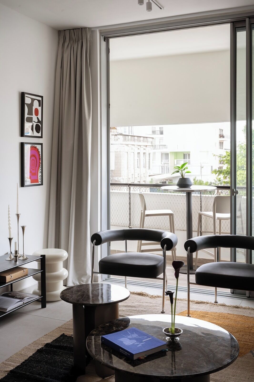 A modern living room with a large window, two black chairs, a round table with a book, and a potted plant on the balcony.
