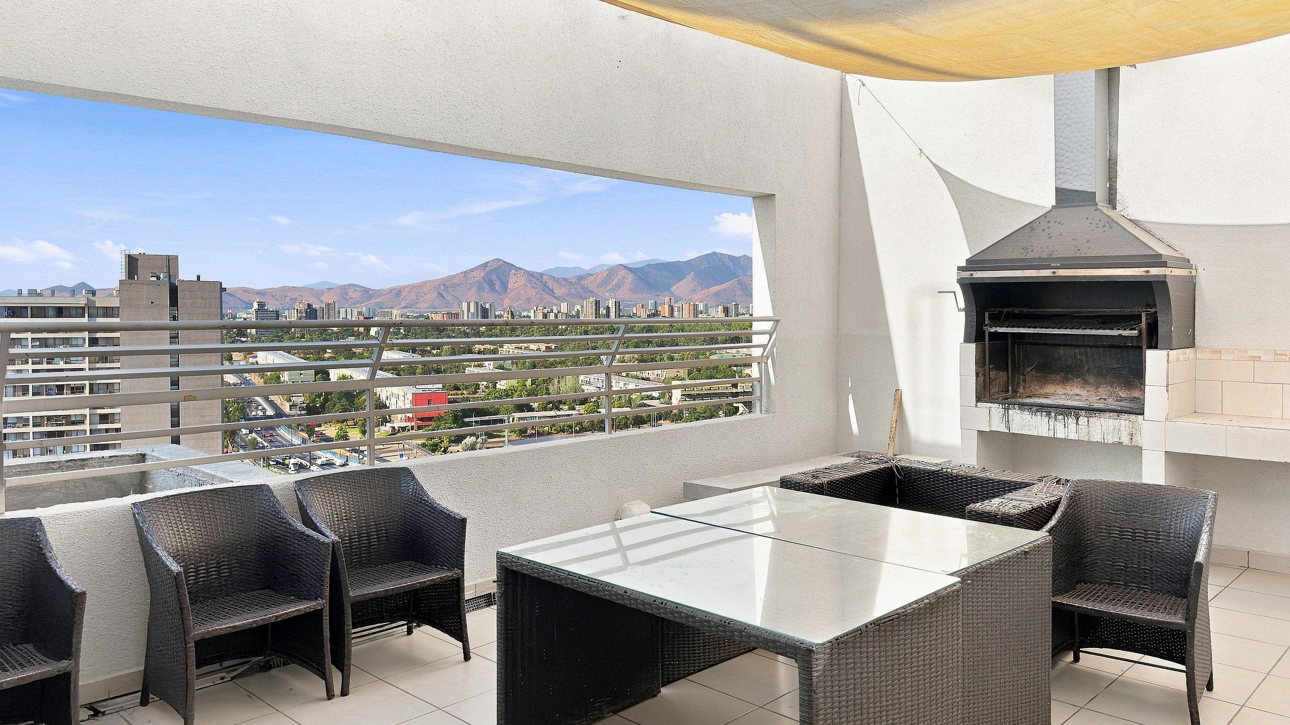 A balcony with a view of a cityscape and mountains, a glass-top table, wicker chairs, and a wood-fired oven.