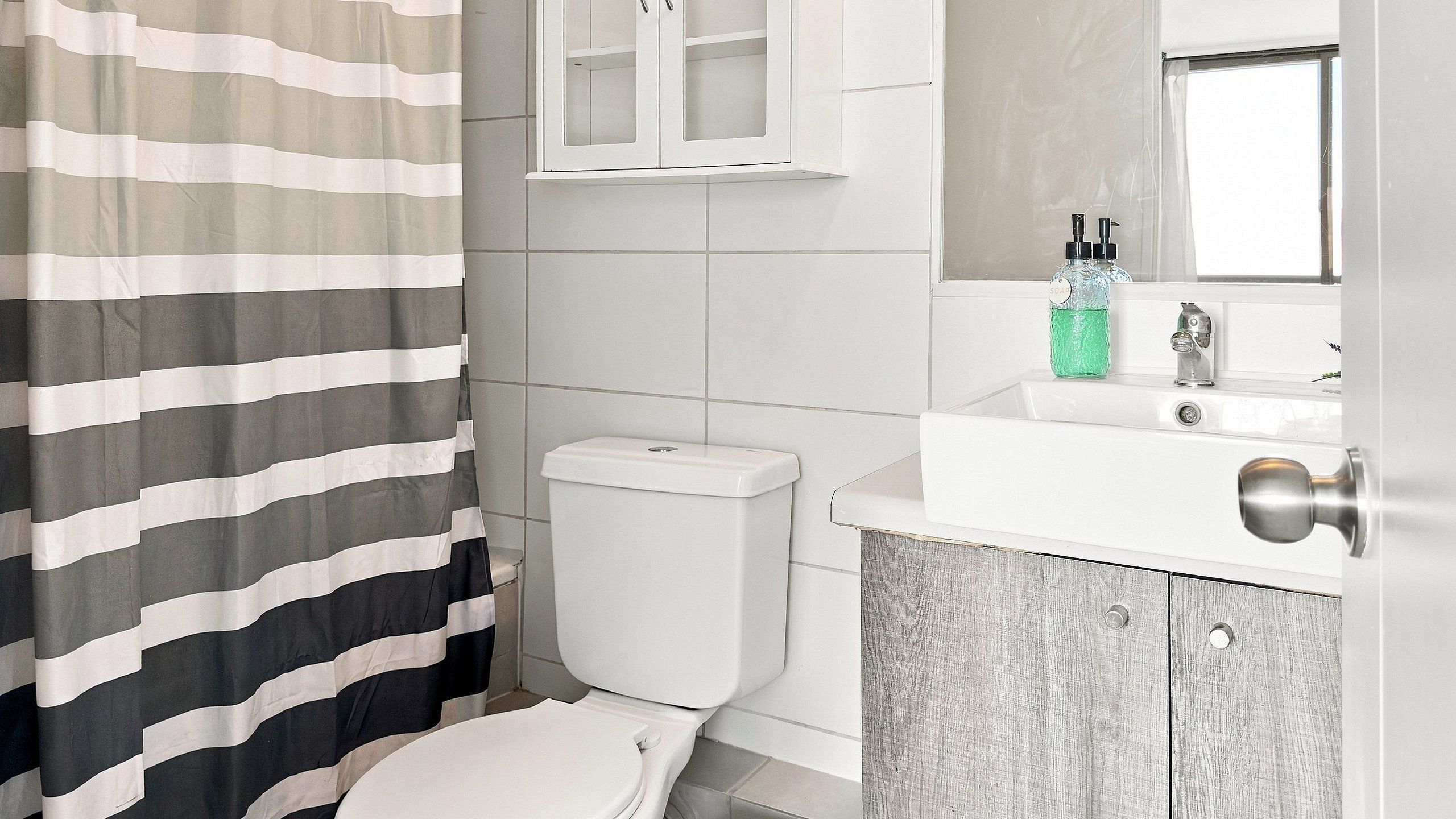 Une salle de bain avec un rideau de douche à rayures, des toilettes blanches, un lavabo blanc avec un distributeur de savon et un miroir au-dessus du lavabo.