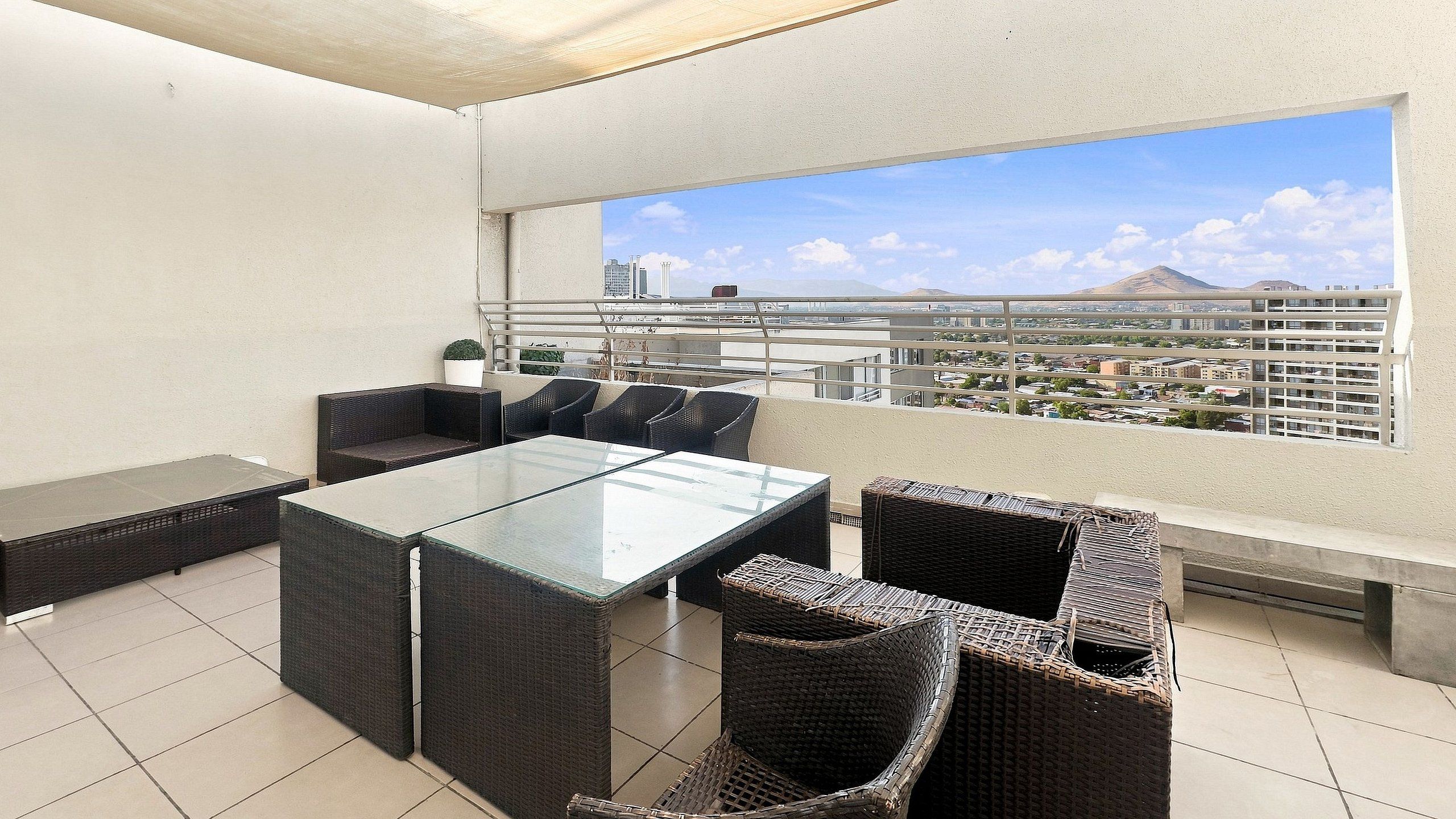 An outdoor balcony with a glass-top table and wicker chairs, overlooking a cityscape with buildings and a mountain.