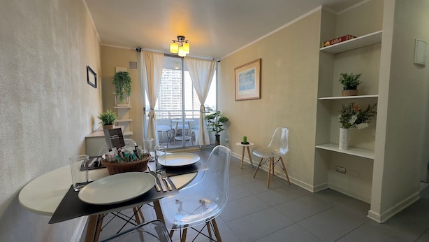 Uma área de refeições com uma mesa posta para dois, cadeiras transparentes, um cesto de petiscos e um vaso de planta.