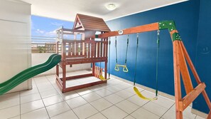 Een kinderspeelplaats met een houten speelhuisje, een glijbaan, schommels en een touwschommel.