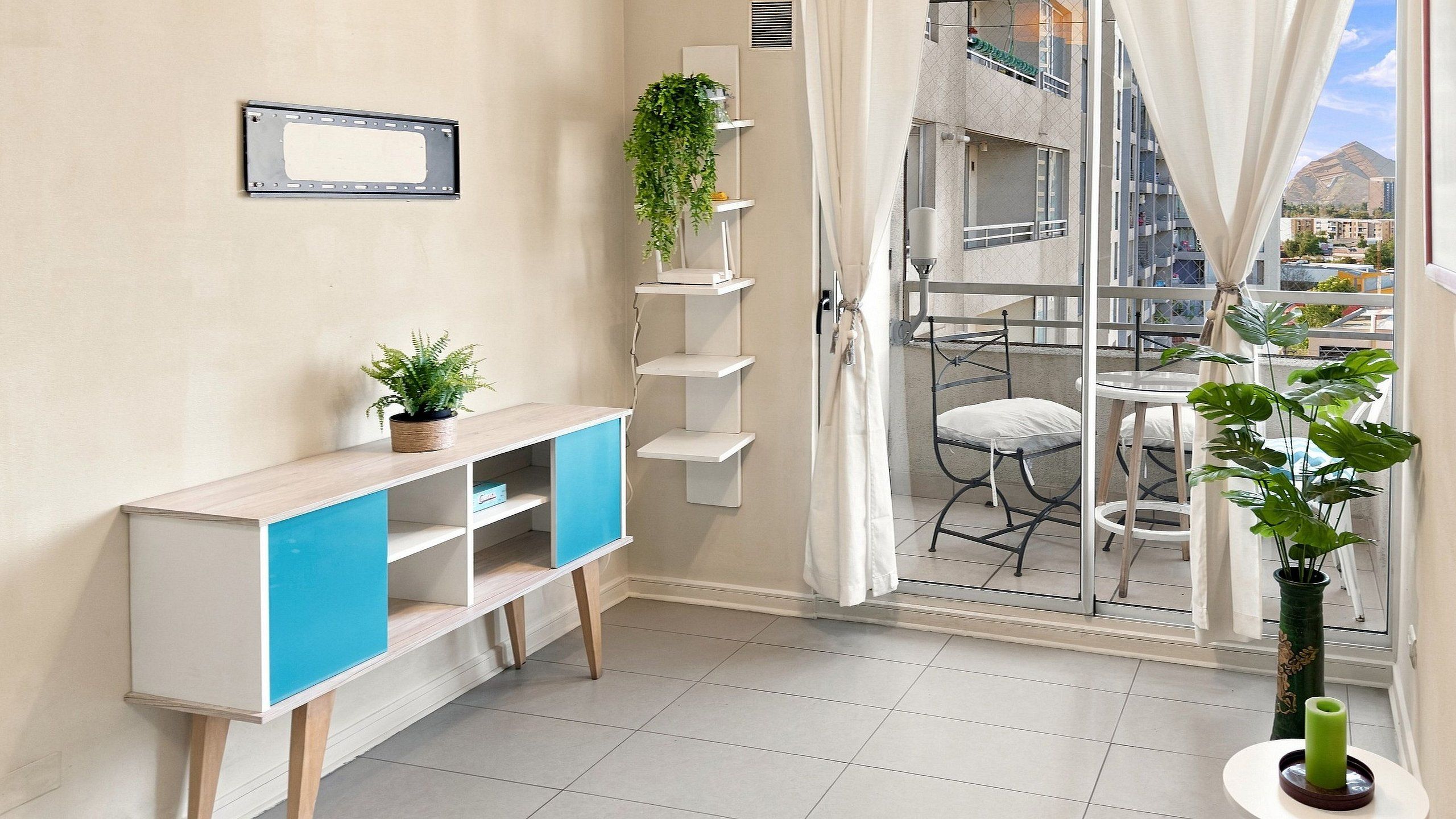 A room with a white console table, a blue cabinet, a potted plant, a shelving unit with plants, a balcony with a chair, and a view of buildings.
