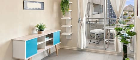 A room with a white console table, a blue cabinet, a potted plant, a shelving unit with plants, a balcony with a chair, and a view of buildings.
