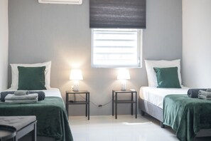 Une chambre d’hôtel avec deux lits, chacun recouvert d’une couvre-lit verte et agrémenté d’oreillers blancs, de part et d’autre desquels se trouvent des tables de chevet avec des lampes. Une fenêtre avec des stores est placée au centre, au-dessus des lits.