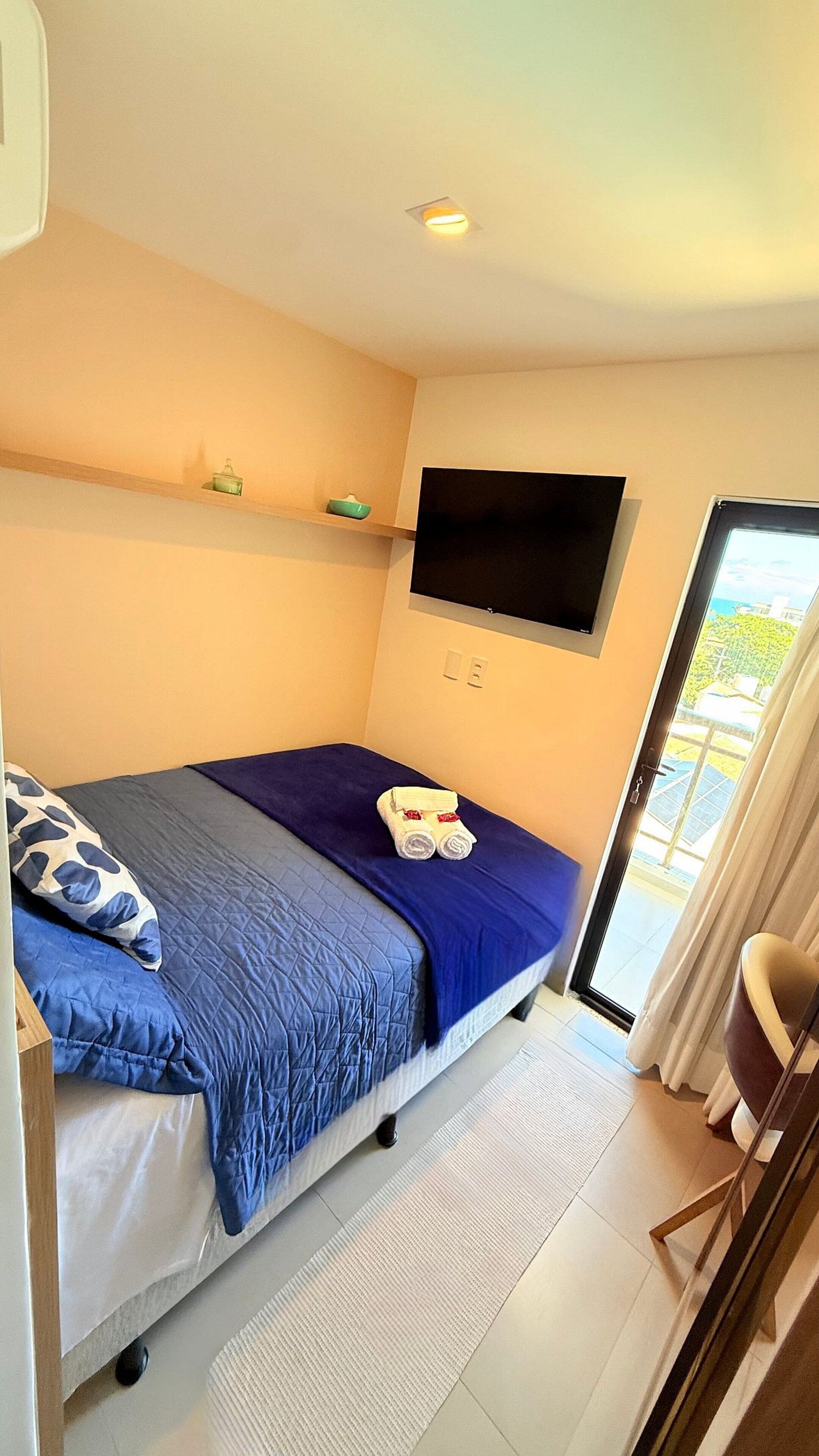 A bedroom with a bed, a TV mounted on the wall, and a view of the outdoors through a window.