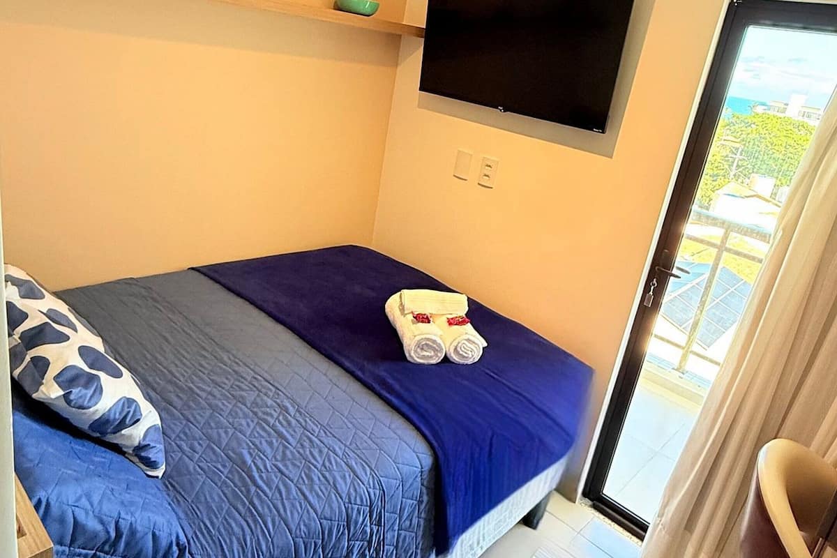 A bedroom with a bed, a TV mounted on the wall, and a view of the outdoors through a window.