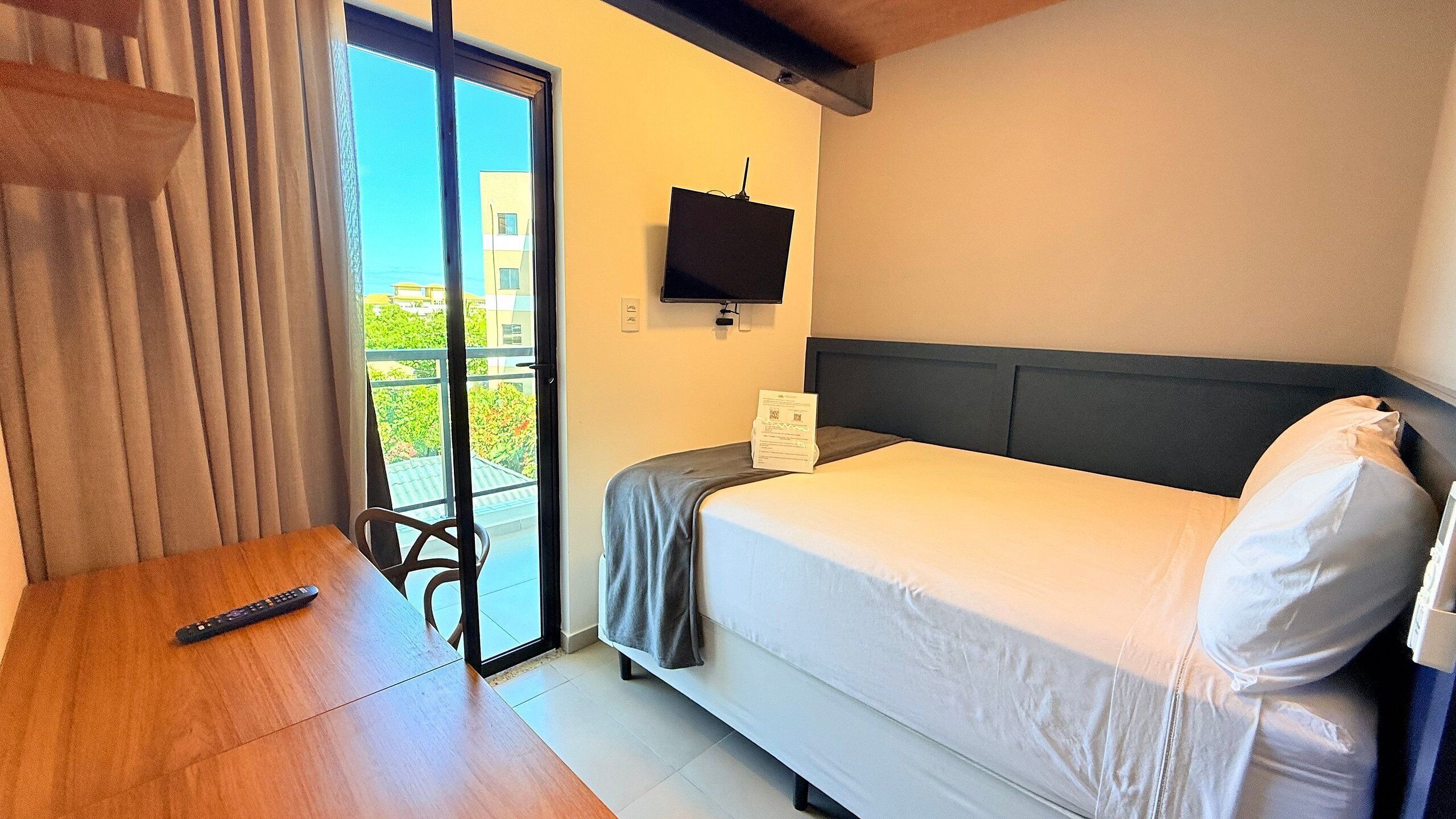 A hotel room with a bed, a TV, a remote control, and a view of greenery outside.
