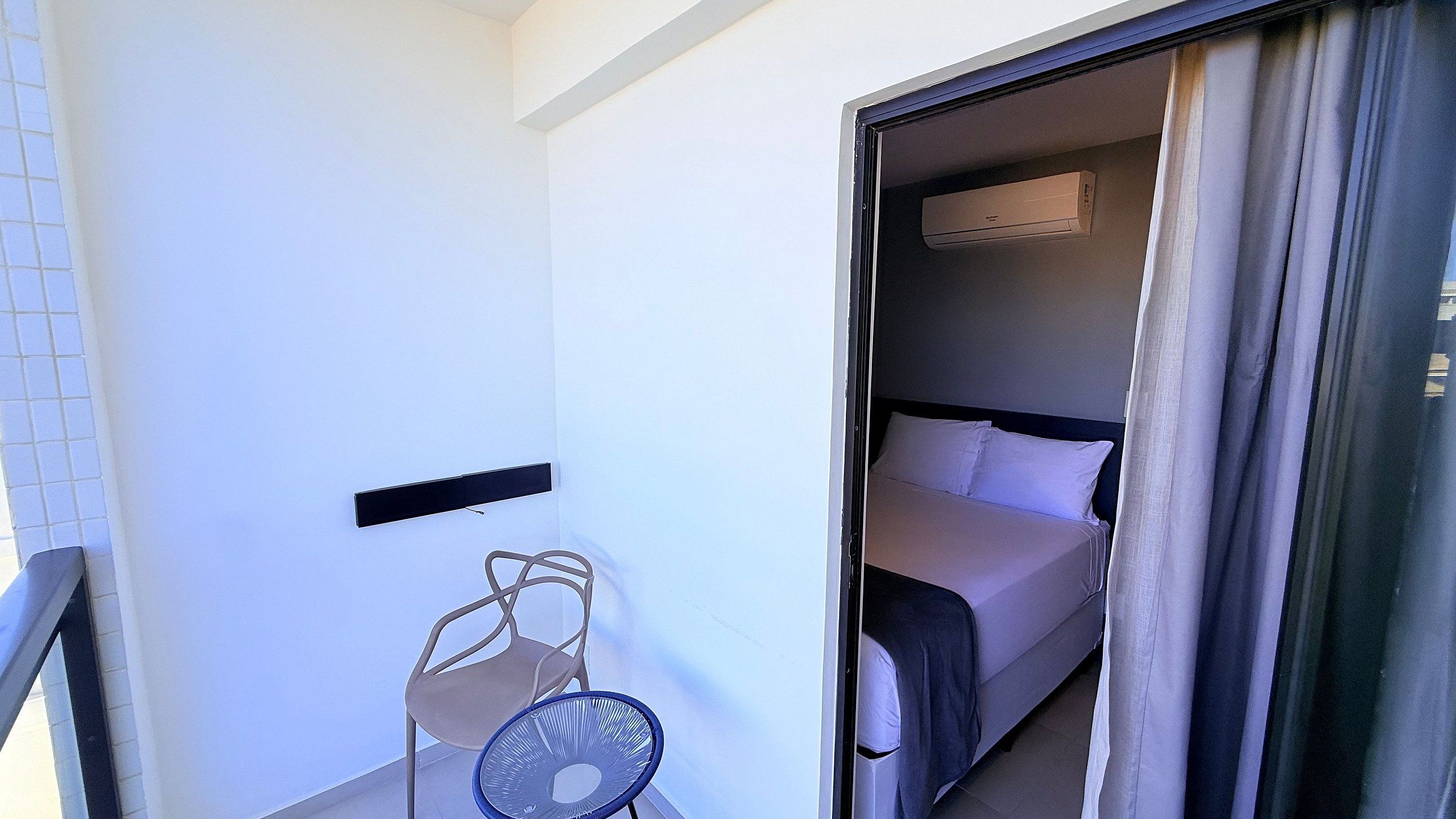 A hotel room with a bed, a wall-mounted air conditioner, a chair, and a window with curtains.