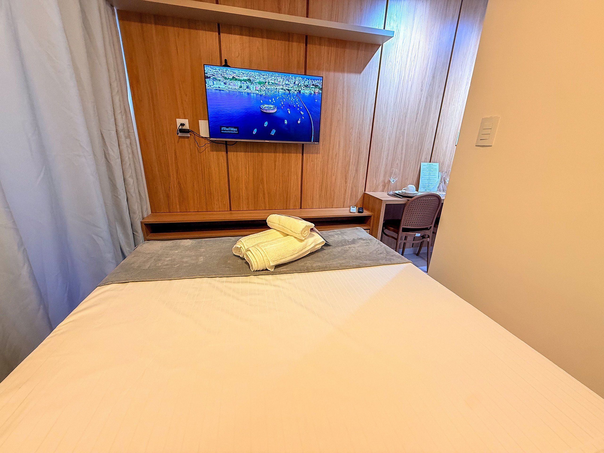 A hotel room with a bed, a TV mounted on the wall, a desk with a chair, and a window with curtains.