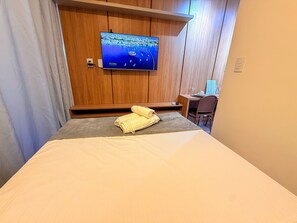 A hotel room with a bed, a TV mounted on the wall, a desk with a chair, and a window with curtains. - Concept Life Stella (Salvador)