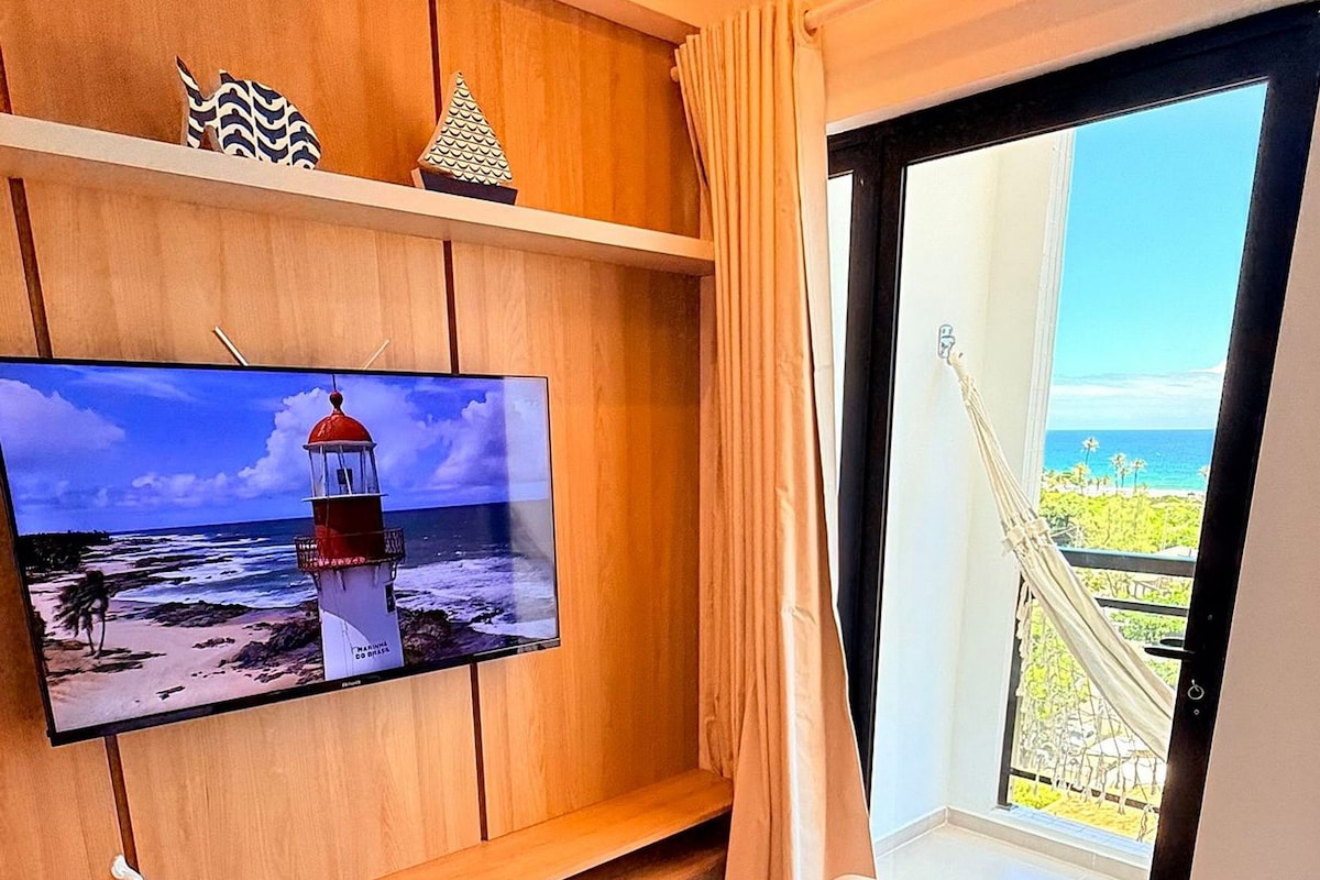 A hotel room with a bed, a TV on a wooden shelf, a view of the sea, and a window with curtains.