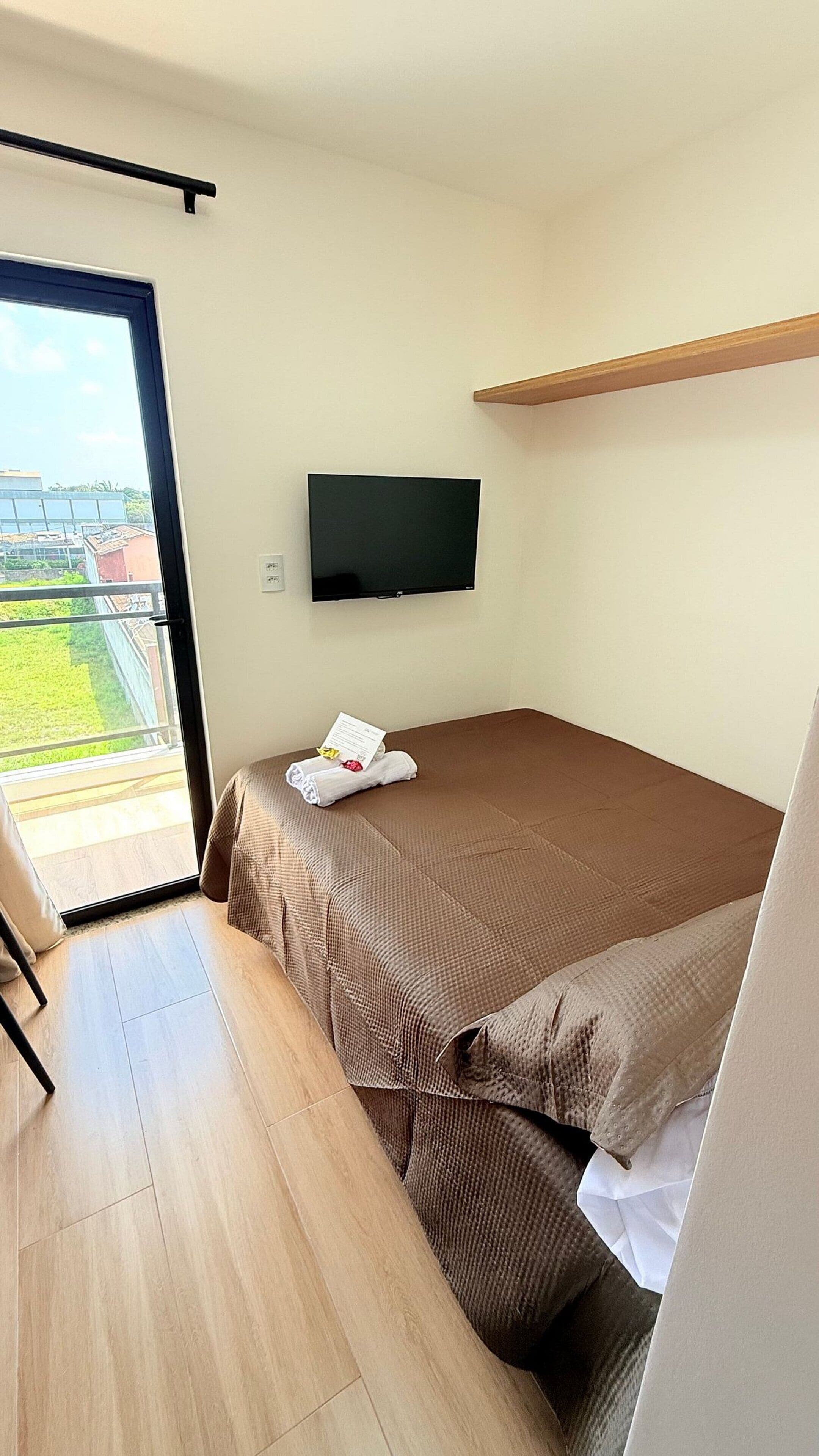 A single bed with a brown bedspread, a flat-screen TV mounted on the wall, a wooden shelf above the bed, and a sliding glass door leading to an outdoor area.