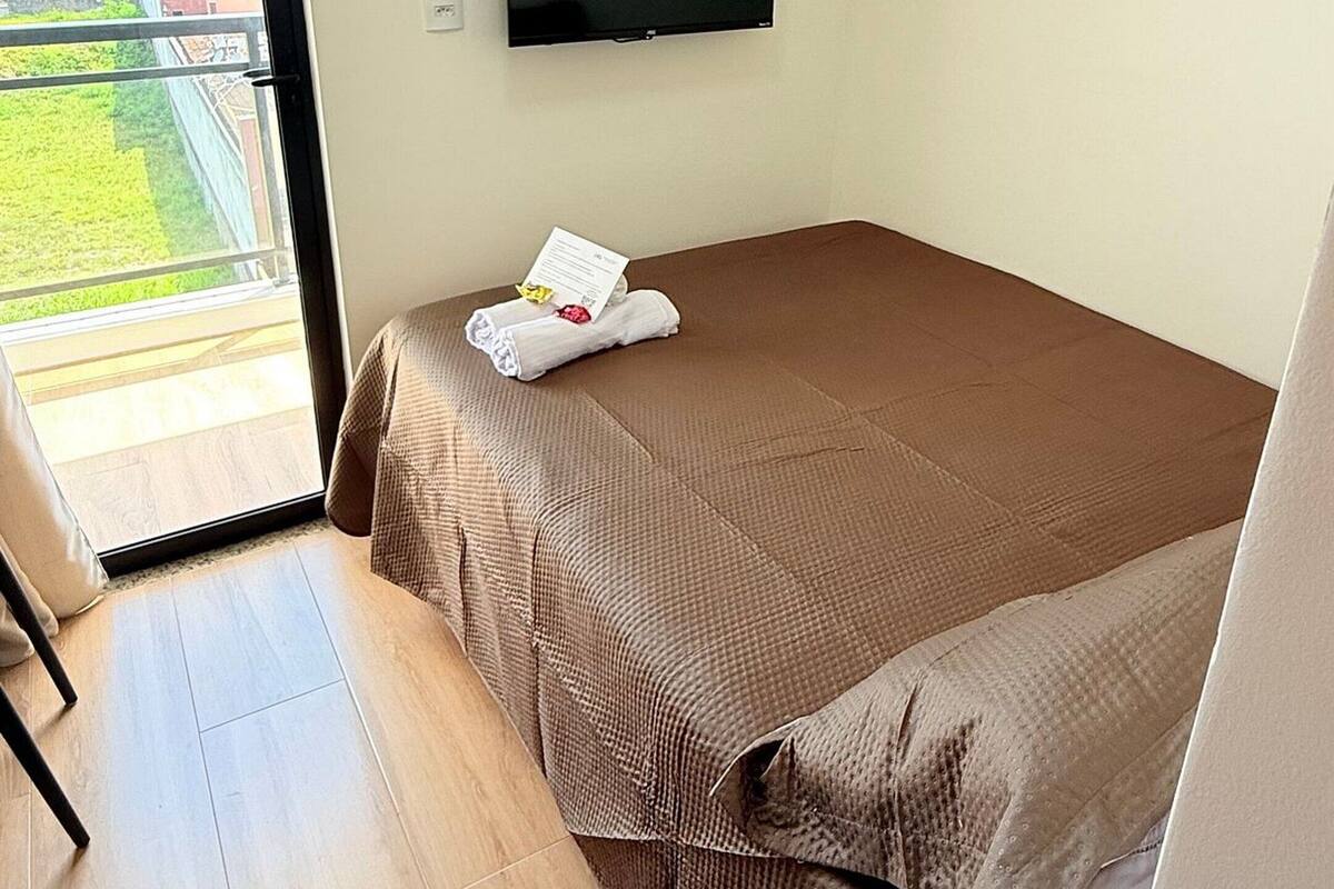 Una cama individual con una colcha marrón, un televisor de pantalla plana montado en la pared, un estante de madera sobre la cama y un ventanal corredero que da a un área exterior.