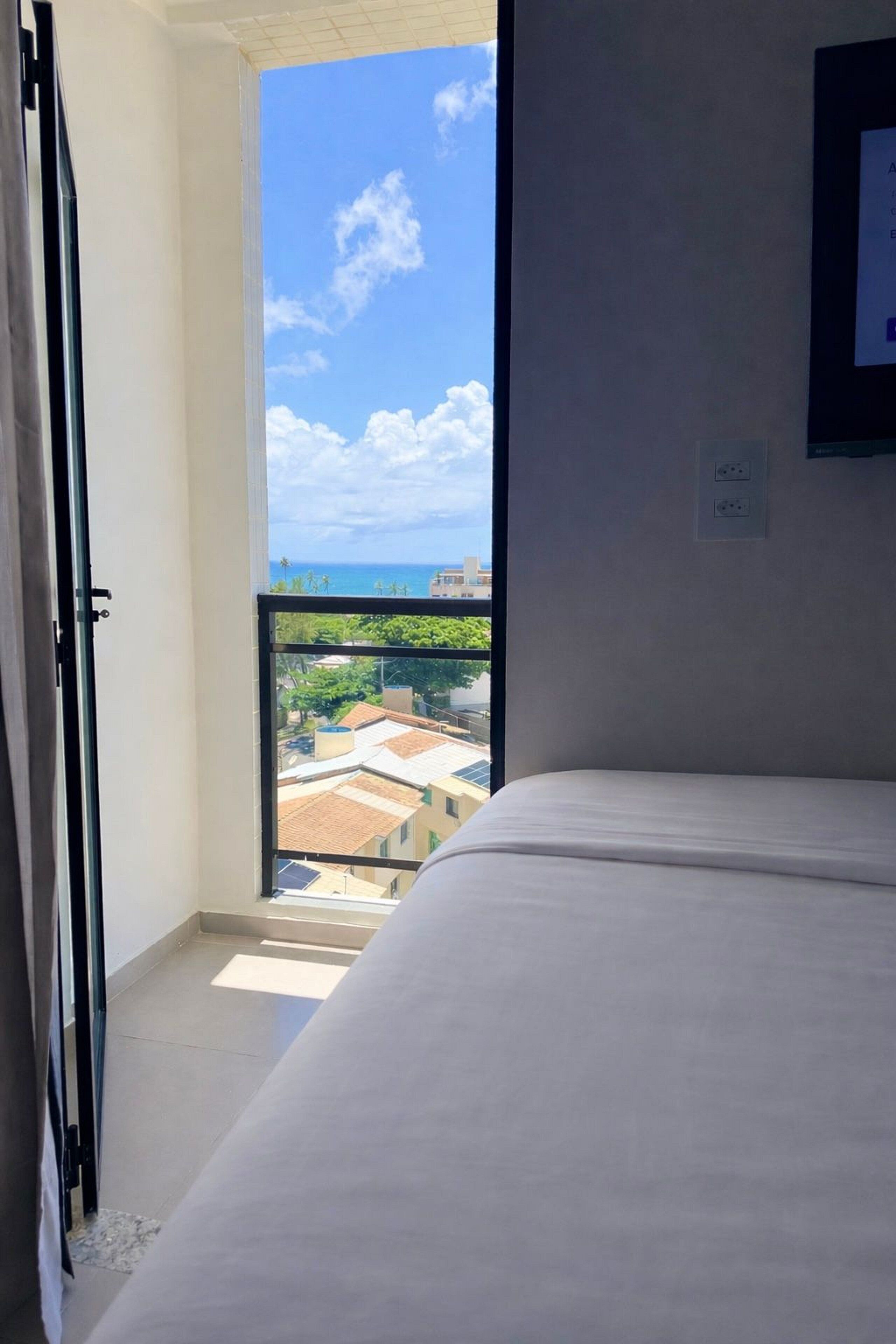 A balcony with a view of buildings and the sea, a bed, and a door.