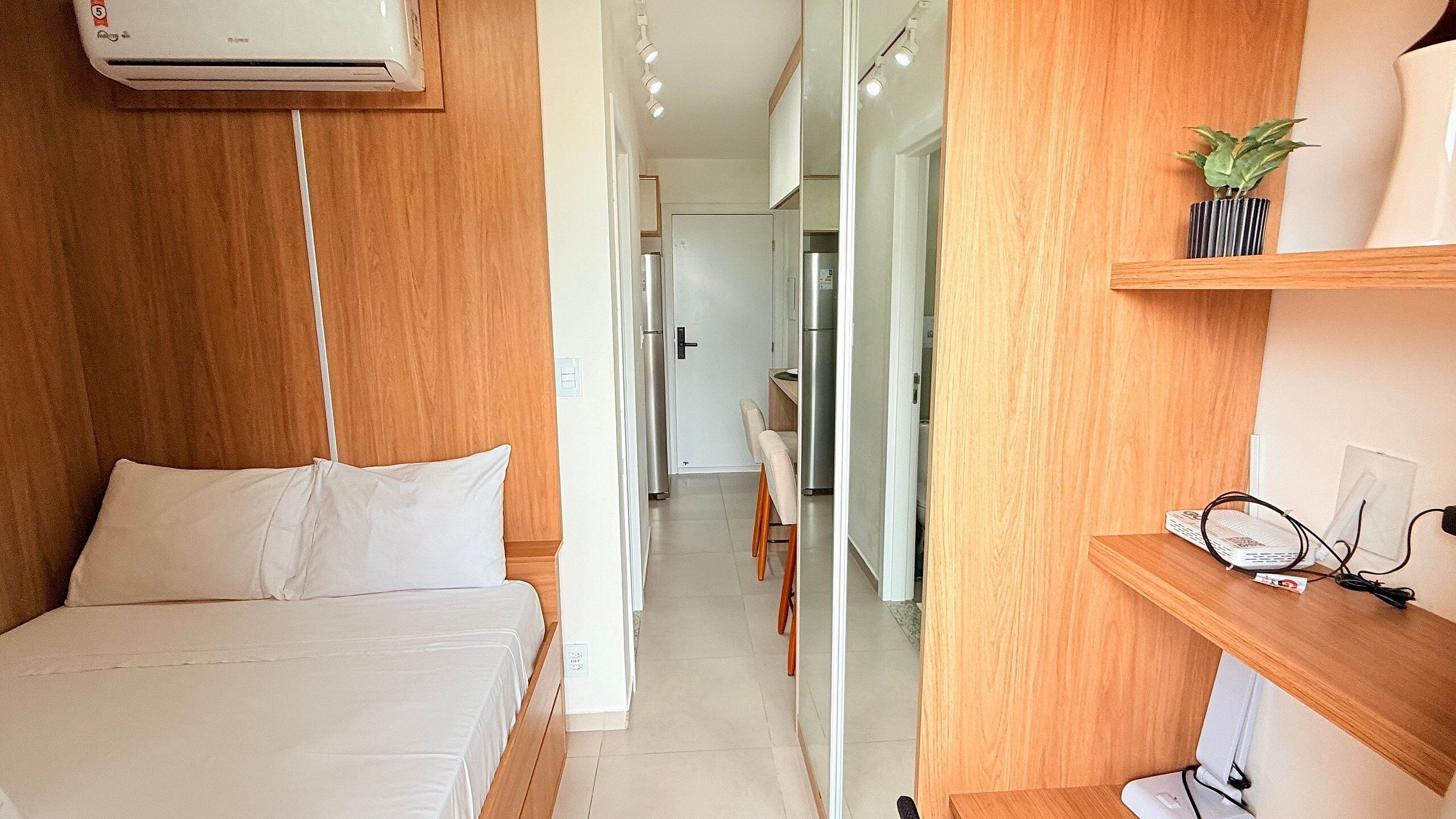 A bedroom with a bed, a desk with a computer, a mirror, and a small plant.