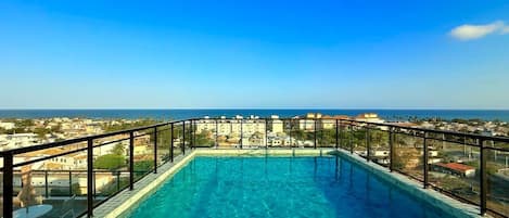 A rooftop pool with clear blue water, surrounded by a black metal railing, overlooking a coastal town.