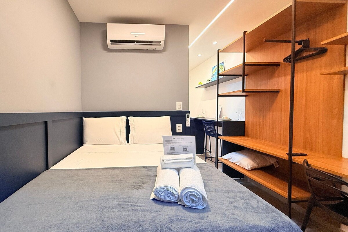 A hotel room with a bed, a desk, a chair, a wardrobe, and an air conditioning unit.