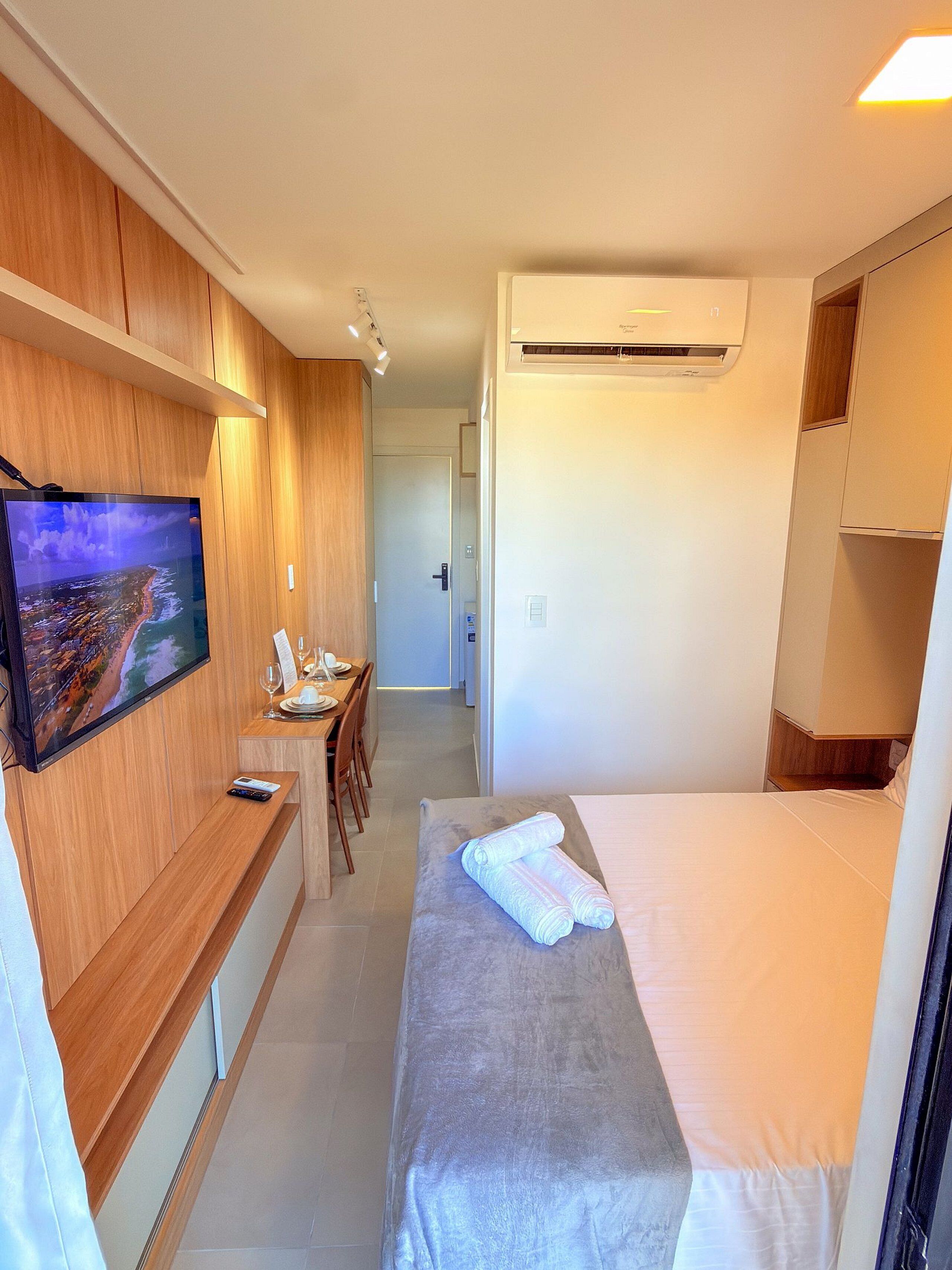 A hotel room with a TV, a desk with a chair, a bed, and an air conditioning unit.