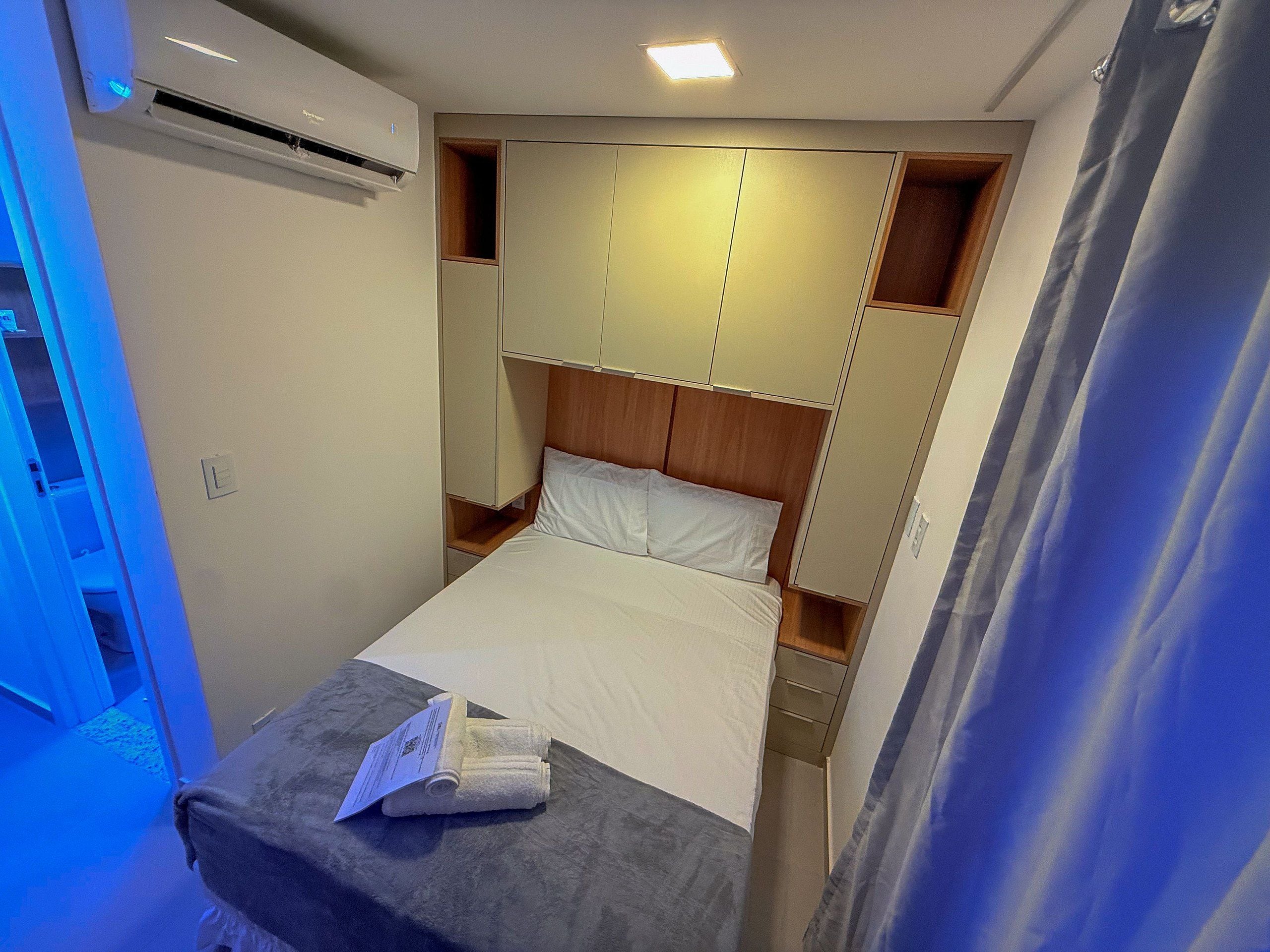 Habitación de hotel compacta con cama, mesita de noche pequeña, armario empotrado y equipo de aire acondicionado.