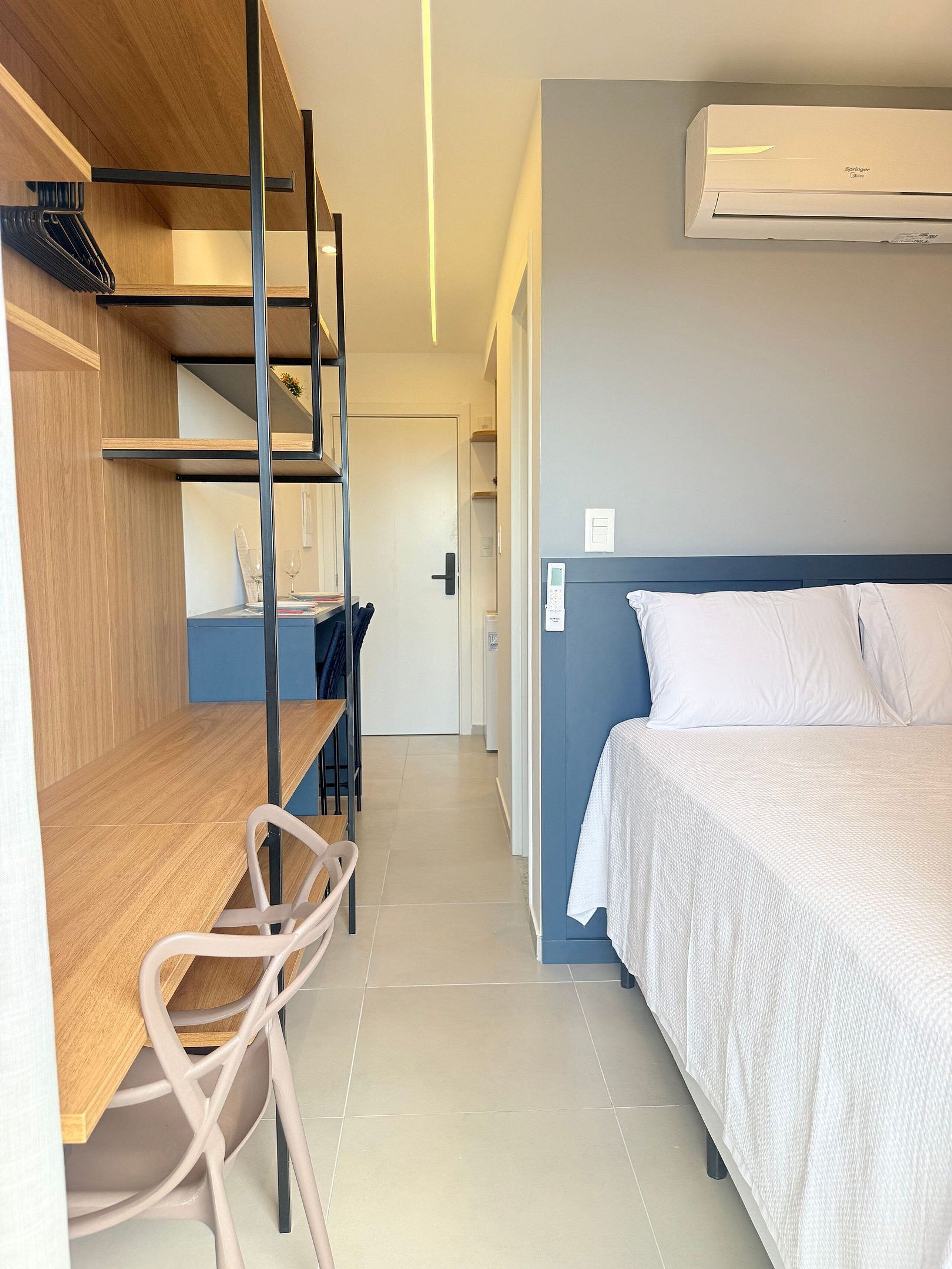 A hotel room with a bed, a desk, a chair, a wardrobe, and an air conditioning unit.