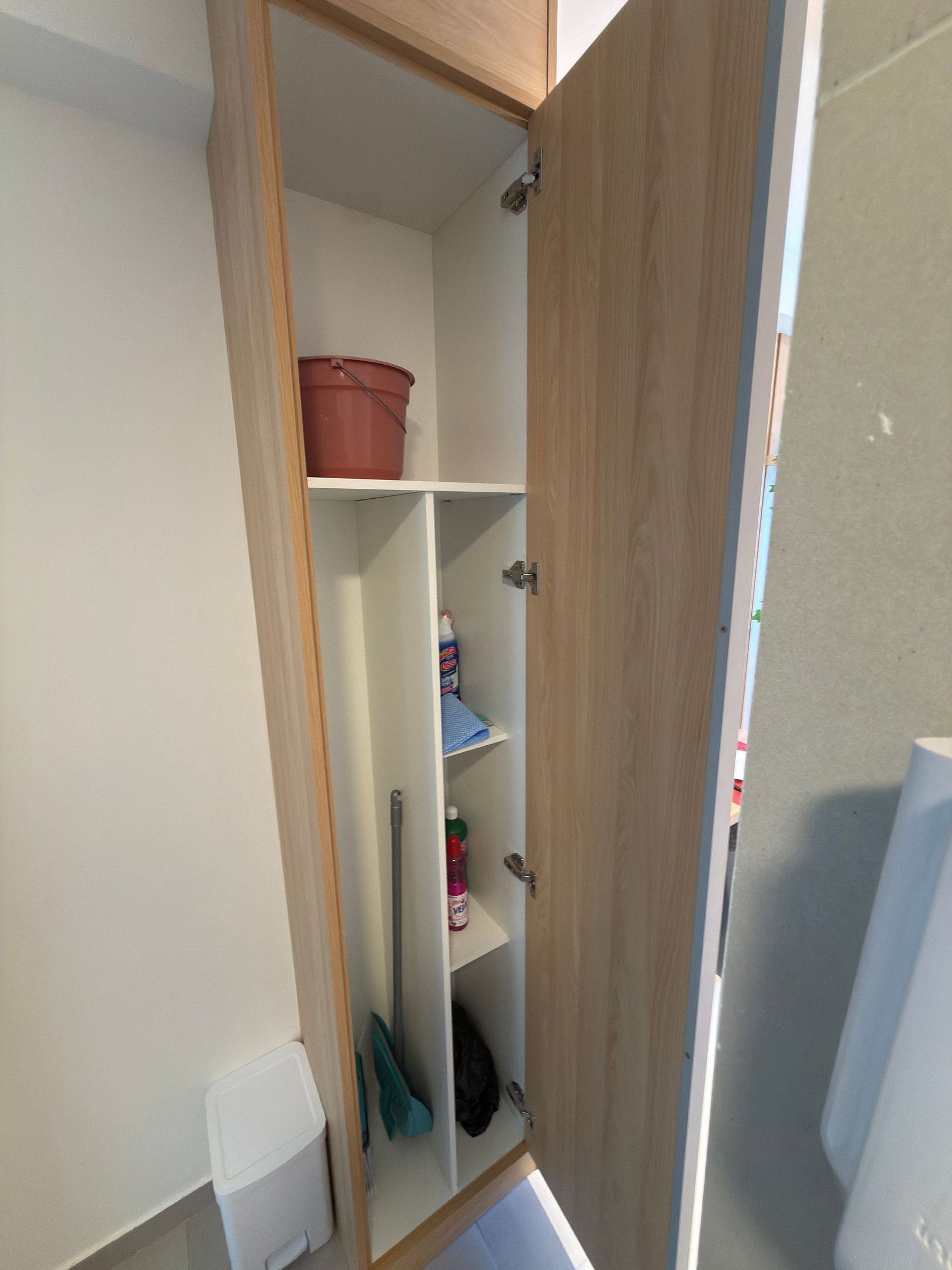 Une armoire entrouverte, avec des étagères contenant des produits de nettoyage et un seau rouge.