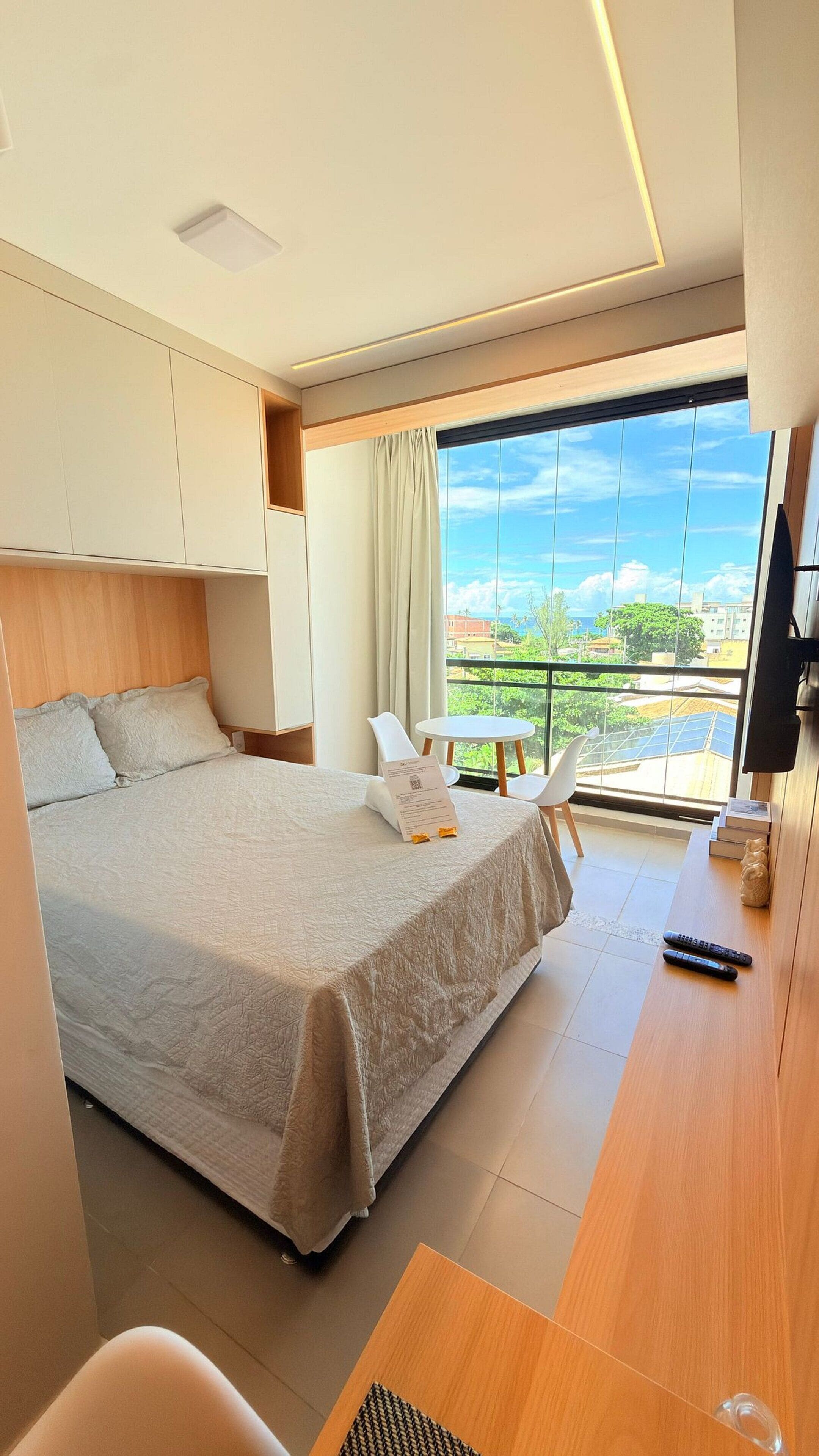 A modern hotel room with a large bed, a desk, a chair, a TV, and a balcony with a city view.