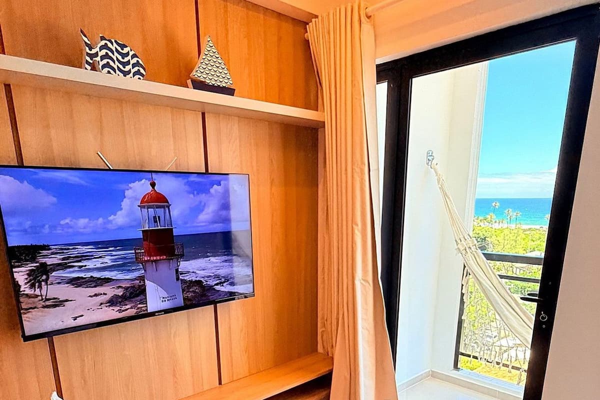 A hotel room with a bed, a TV on a wooden shelf, a view of the sea, and a window with curtains.