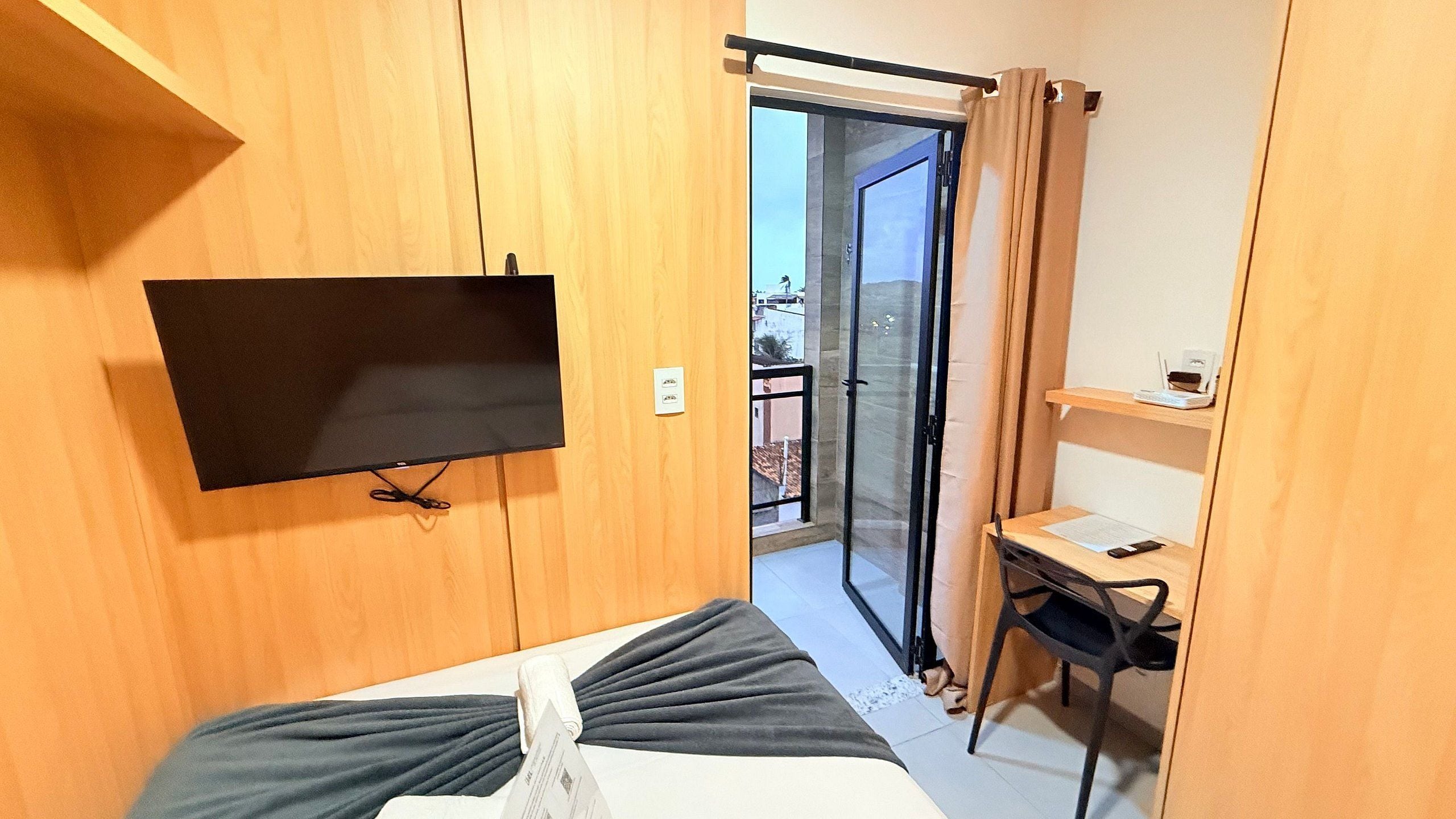 Una habitación con una cama, un televisor montado en la pared, un escritorio con una silla y un ventanal corredero que da a un espacio exterior.