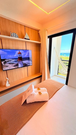 A hotel room with a bed, a TV on a wooden shelf, a view of the sea, and a window with curtains. - Concept Life Stella (Salvador)