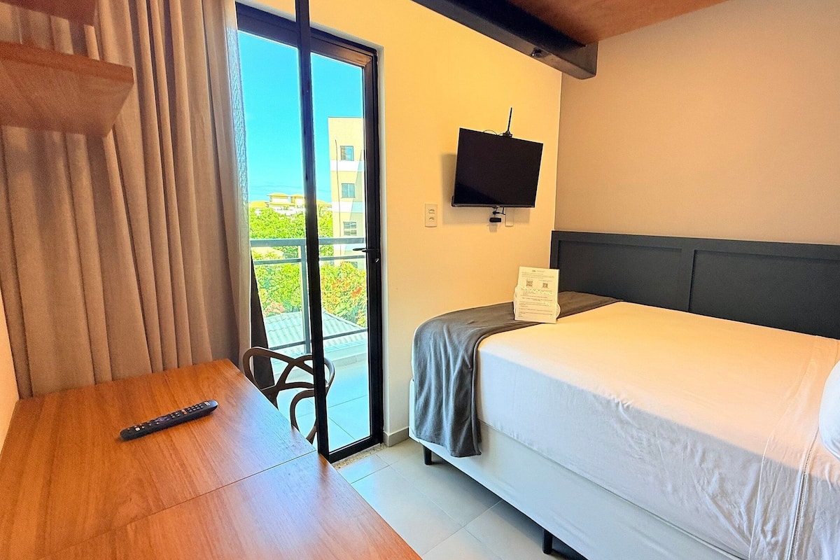 A hotel room with a bed, a TV, a remote control, and a view of greenery outside.