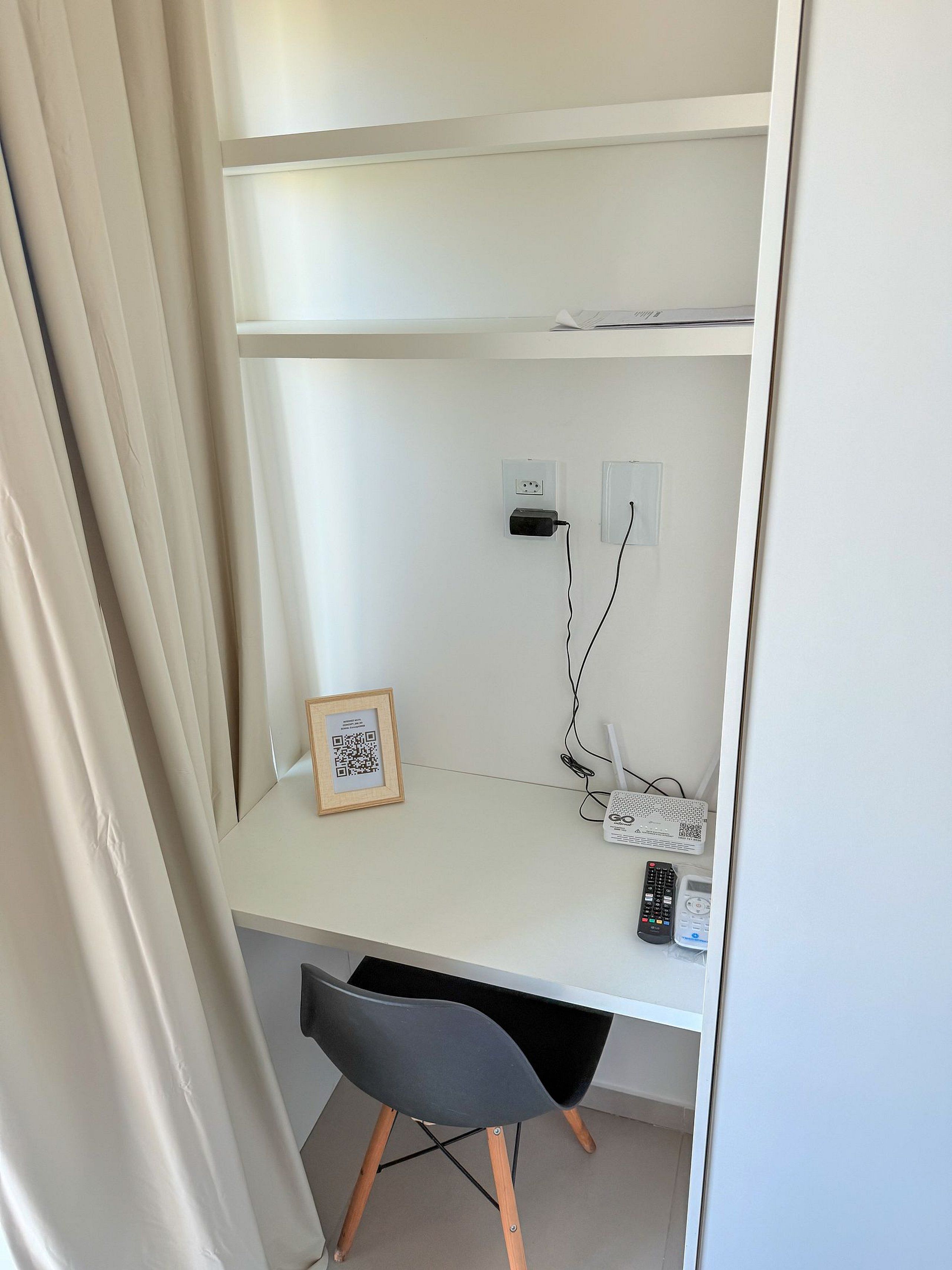 Un escritorio blanco con una silla negra, un cuadro enmarcado y un router en la pared.