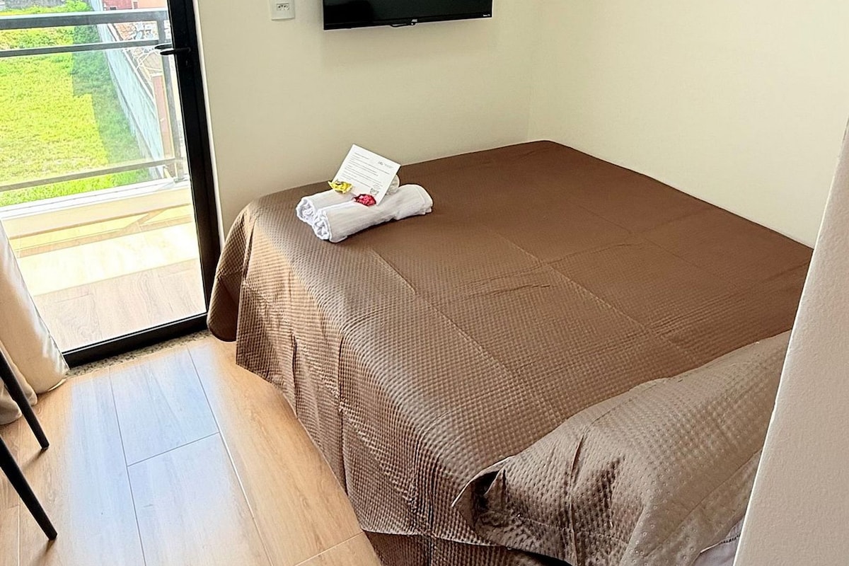 A single bed with a brown bedspread, a flat-screen TV mounted on the wall, a wooden shelf above the bed, and a sliding glass door leading to an outdoor area.