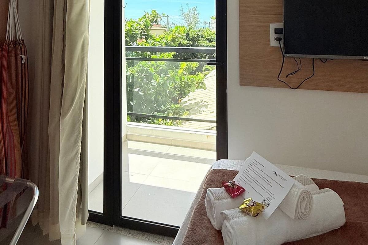 A hotel room with a bed, a TV mounted on the wall, a window with a view of greenery, and a door leading to an outdoor balcony.