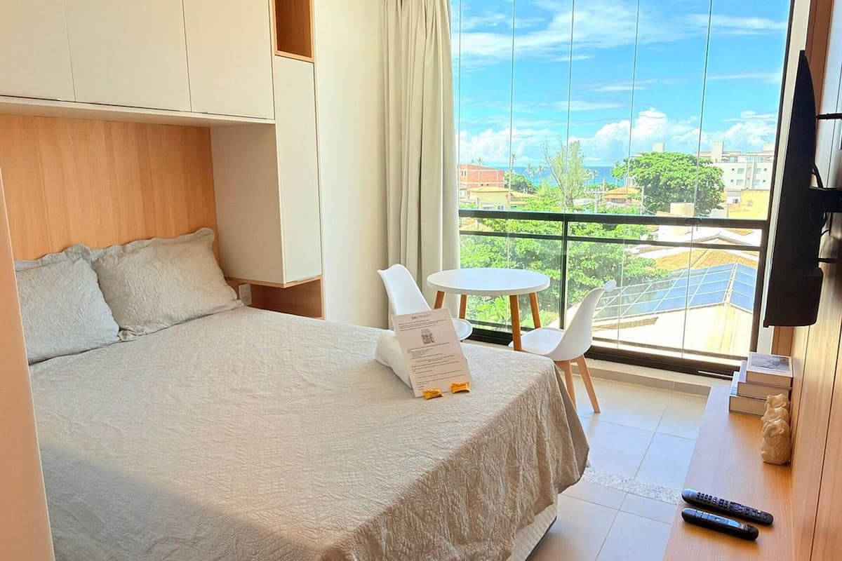 A modern hotel room with a large bed, a desk, a chair, a TV, and a balcony with a city view.