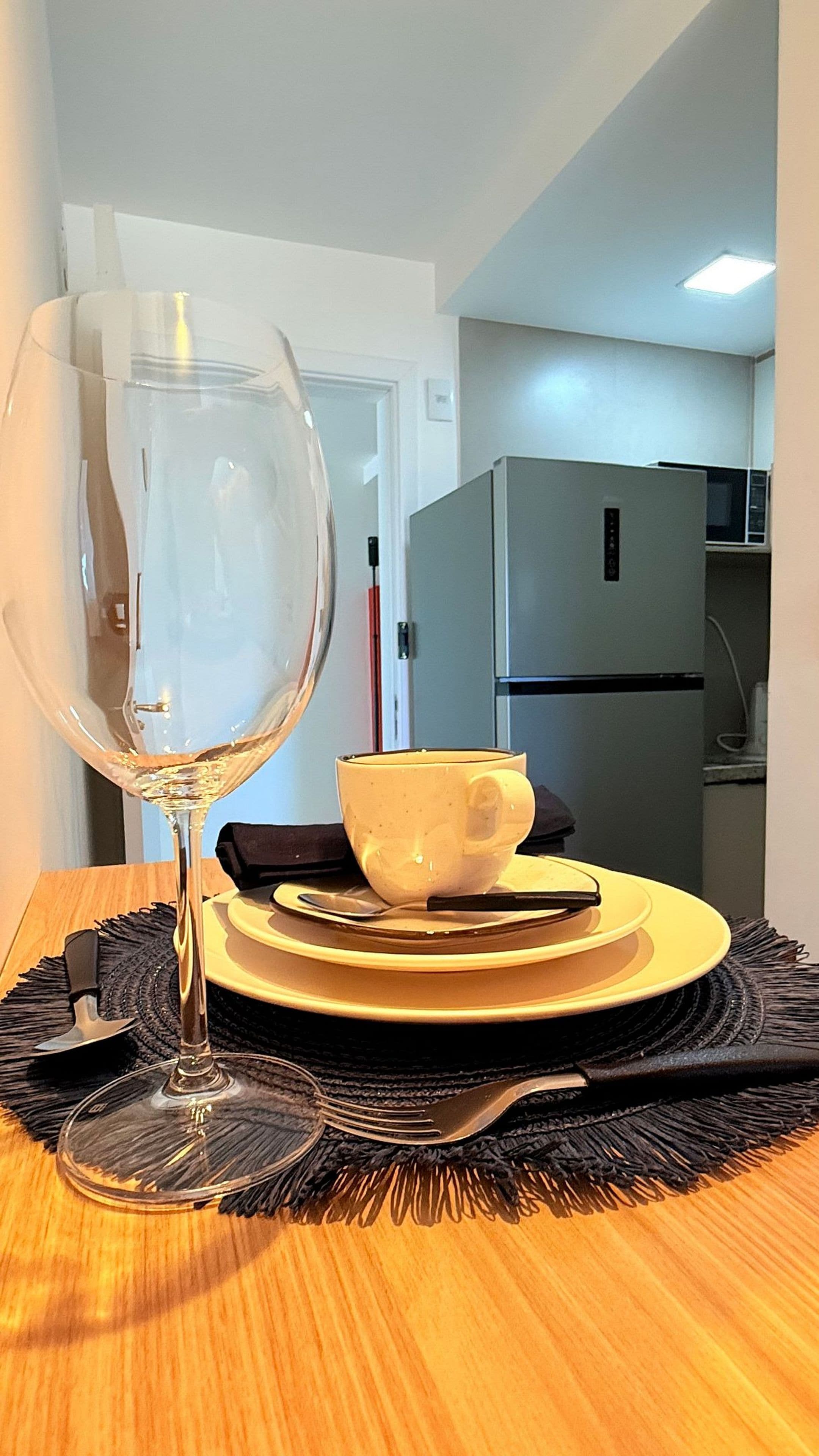A dining table with a clear glass, a yellow bowl, and plates. A microwave and refrigerator are in the background.