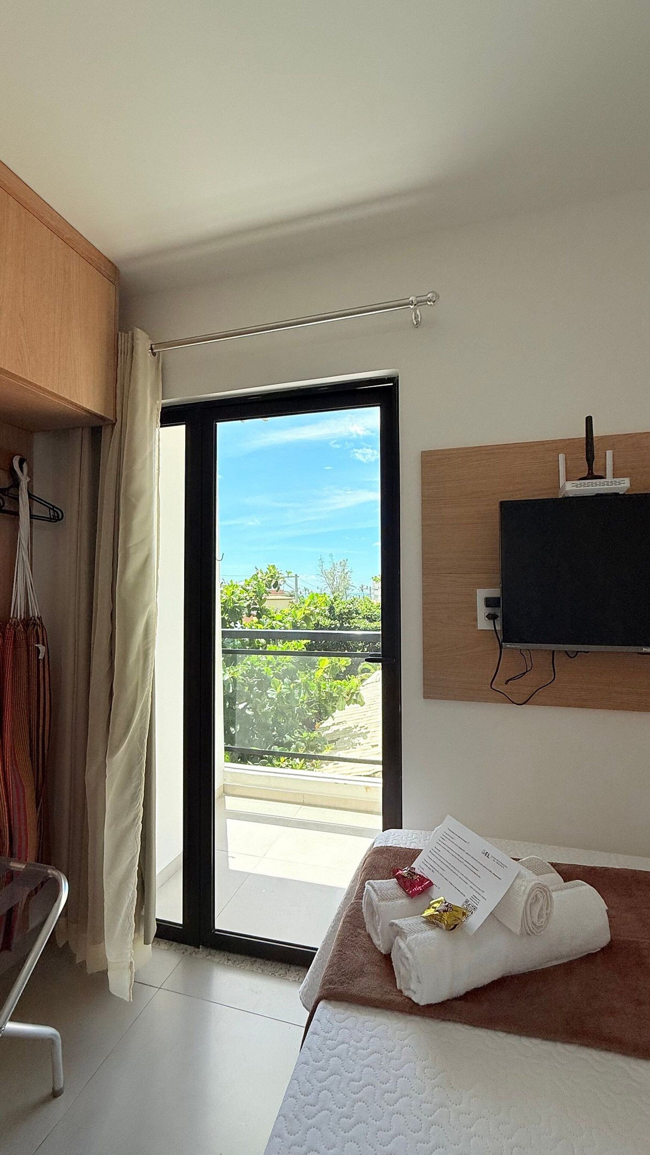 A hotel room with a bed, a TV mounted on the wall, a window with a view of greenery, and a door leading to an outdoor balcony.