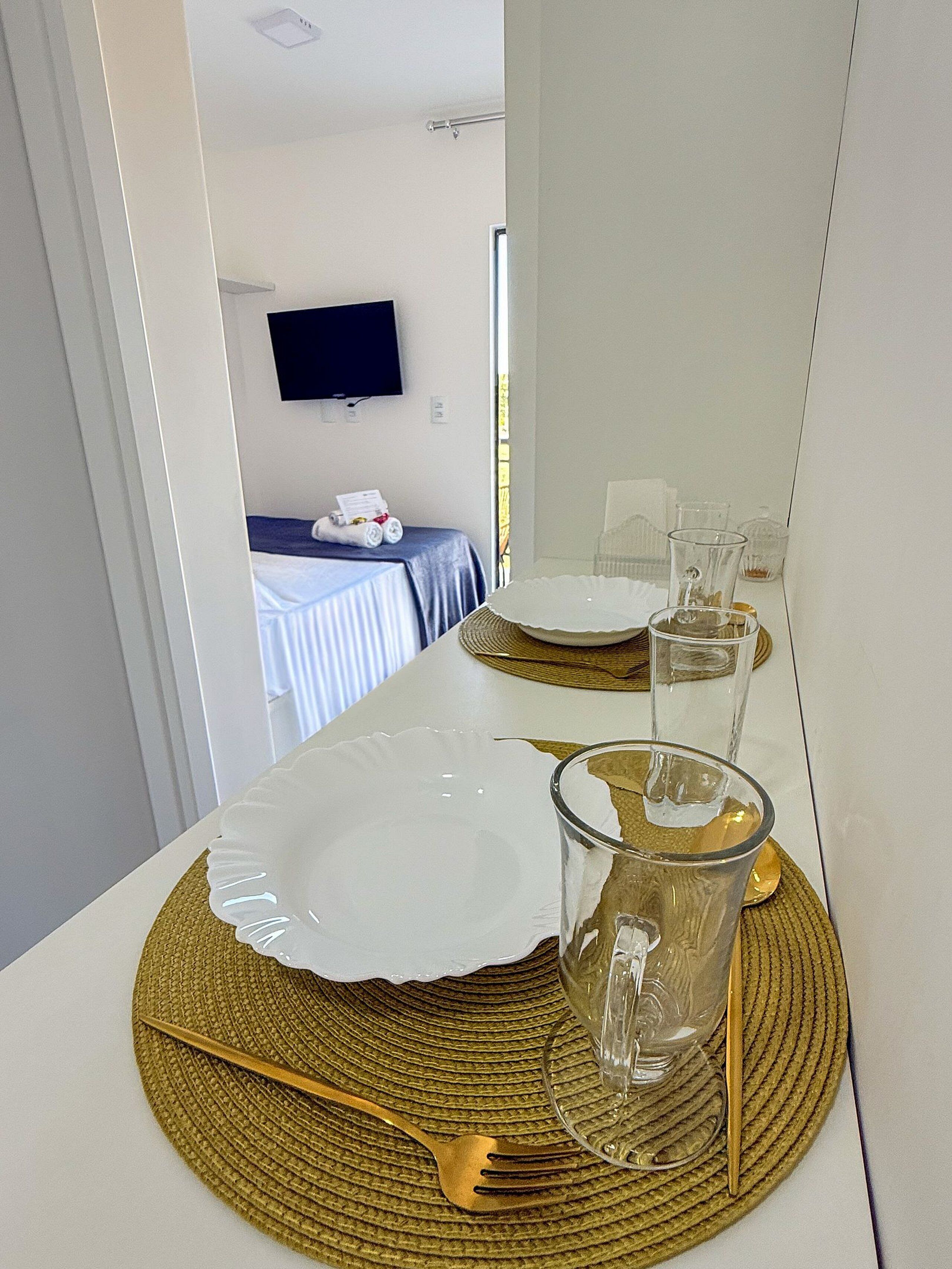 A dining area with a table set for two, a television mounted on the wall, and a window with curtains.