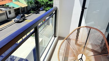 Un balcón con barandilla de metal, una silla de mimbre y vista a una calle con coches y edificios.