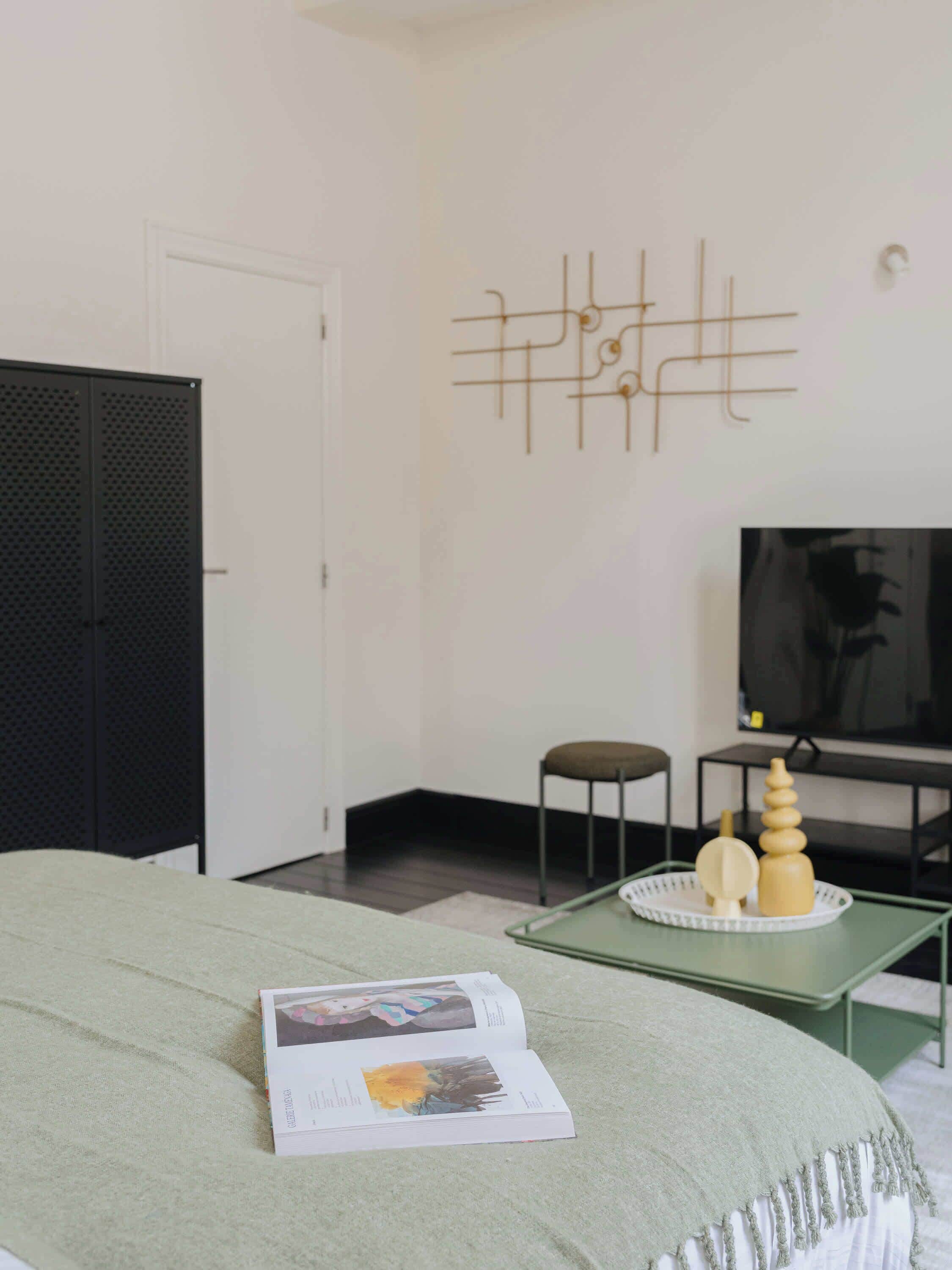 A modern bedroom with a green bedspread, a book on the bed, a TV on a stand, and a decorative wall piece.