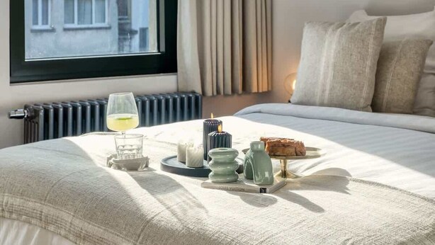 Ein Hotelzimmer mit großem Fenster, einem Bett mit weißer Bettwäsche, einem Nachttisch mit einem Glas Wasser, einer Kerze und einem Buch mit dem Titel „LOUIS VUITTON“.