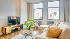 Ein modernes Wohnzimmer mit einem Flachbildfernseher, einer Holz-Unterhaltungseinheit, einem runden Couchtisch mit Blumenvase, einem beigen Sofa und großen Fenstern mit Blick nach draußen.