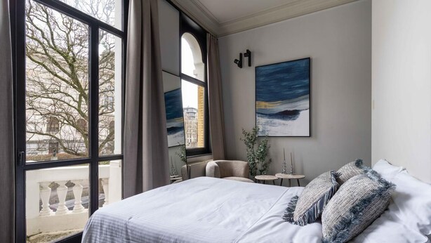A bedroom with a large window, a bed with white linens, a small round table, and a painting on the wall. - Elegant Balcony Suite (Antwerpen)