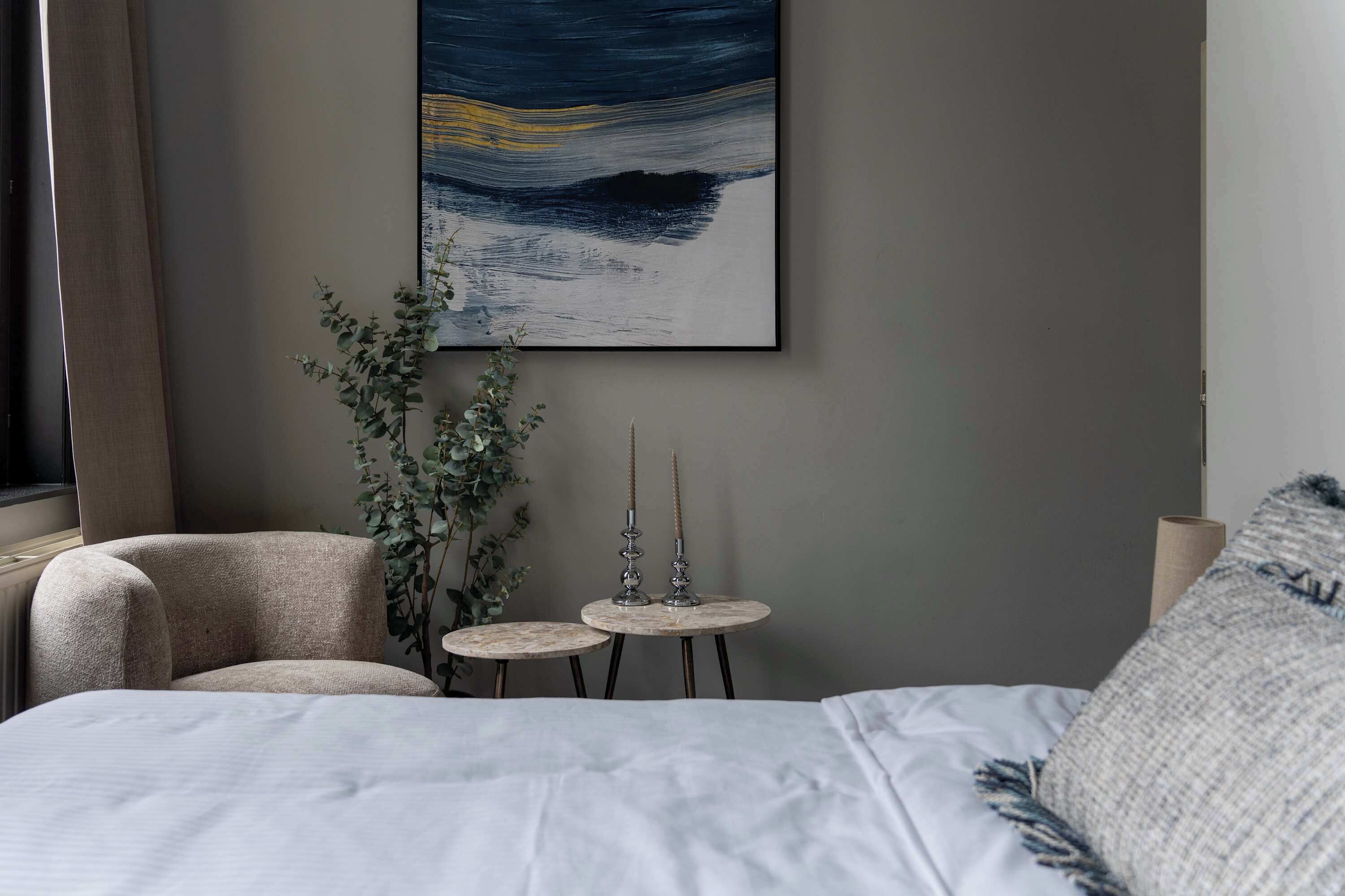 A bedroom with a bed, a chair, two small tables, a plant, and a wall-mounted painting.