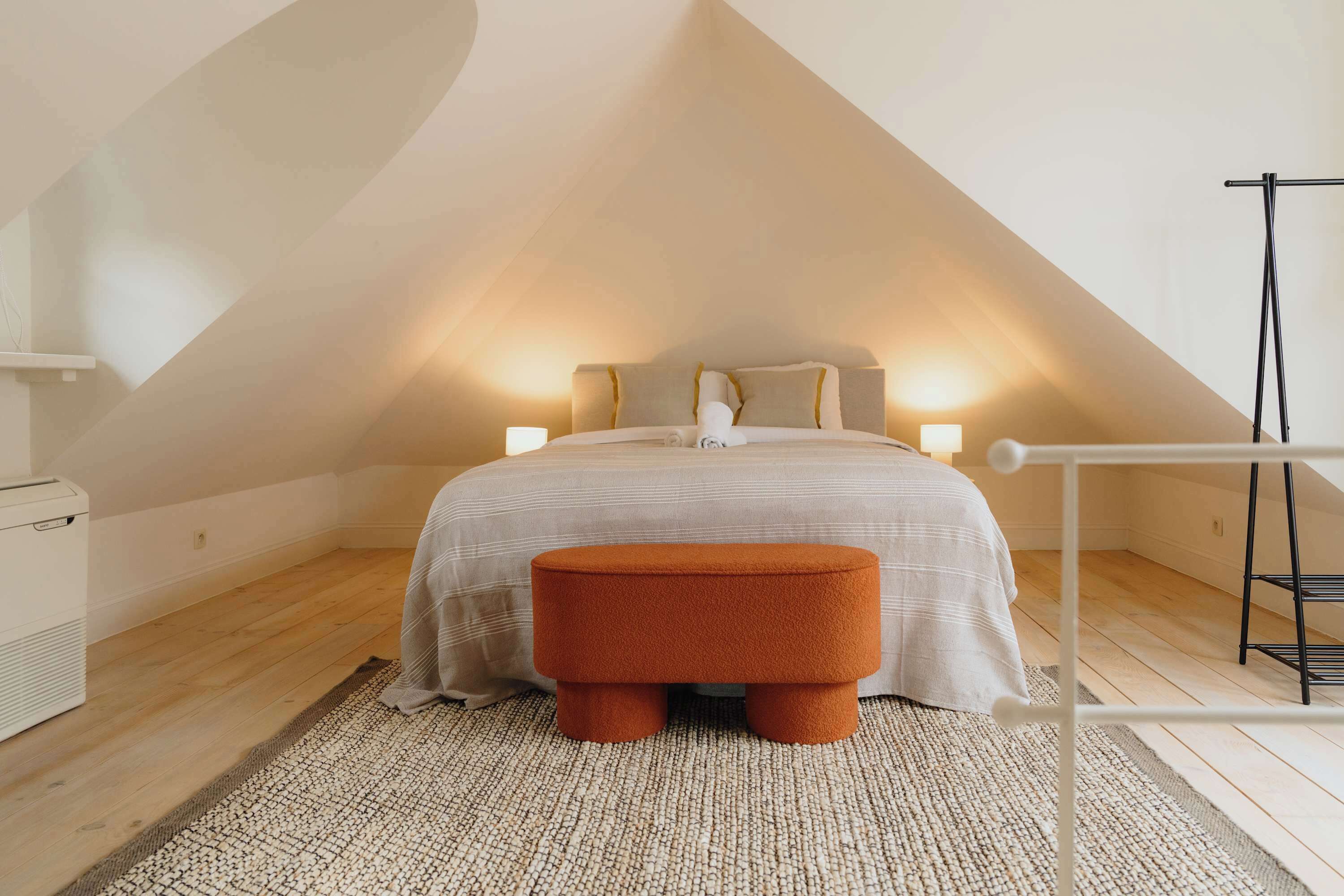 Een slaapkamer met een bed, bedlampjes, een houten vloer, een vloerkleed en een klein oranje pouf.