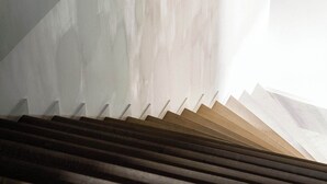Un escalier avec des marches en bois menant vers le haut, entouré d’un mur sobre, une source de lumière créant un effet d’ombre.